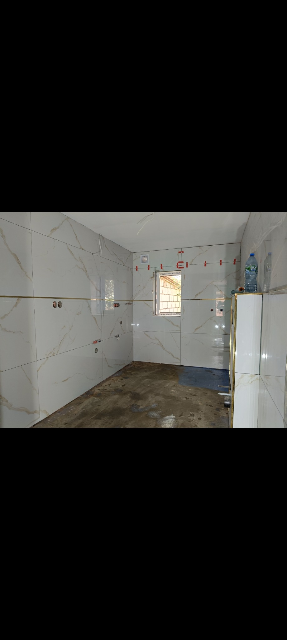 Ściany łazienki w trakcie układania płytek imitujących marmur, z widocznymi krzyżykami dystansowymi i poziomą listwą dekoracyjną. Przy oknie butelka wody.