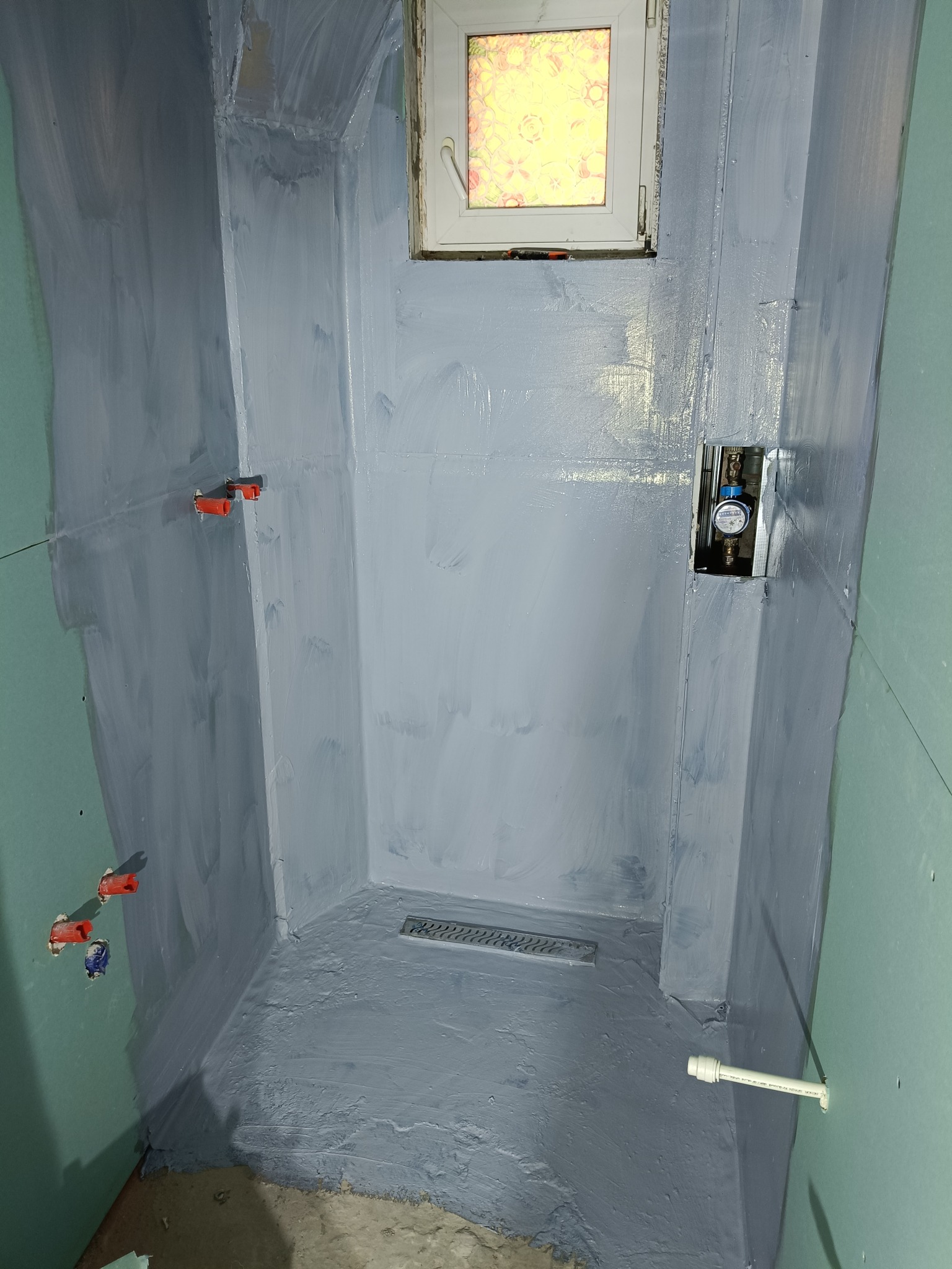 Hydroizolacja w szarej tonacji na ścianach i podłodze kabiny prysznicowej z odpływem liniowym. Widoczne przyłącza wodne i okno.