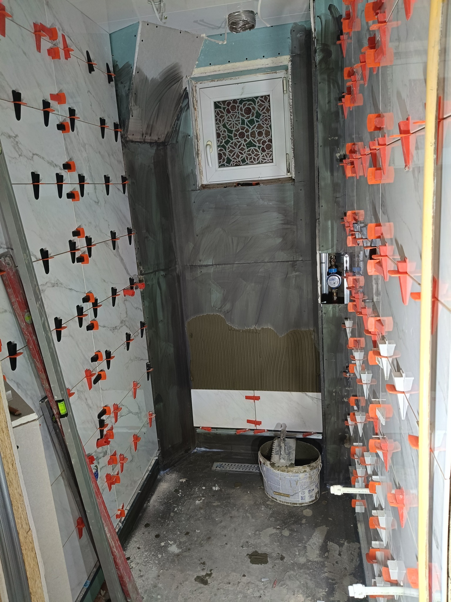 Wnętrze łazienki w trakcie prac glazurniczych: białe płytki imitujące marmur na ścianach, pomarańczowe klipsy poziomujące, widoczny fragment okna i narzędzia glazurnicze.