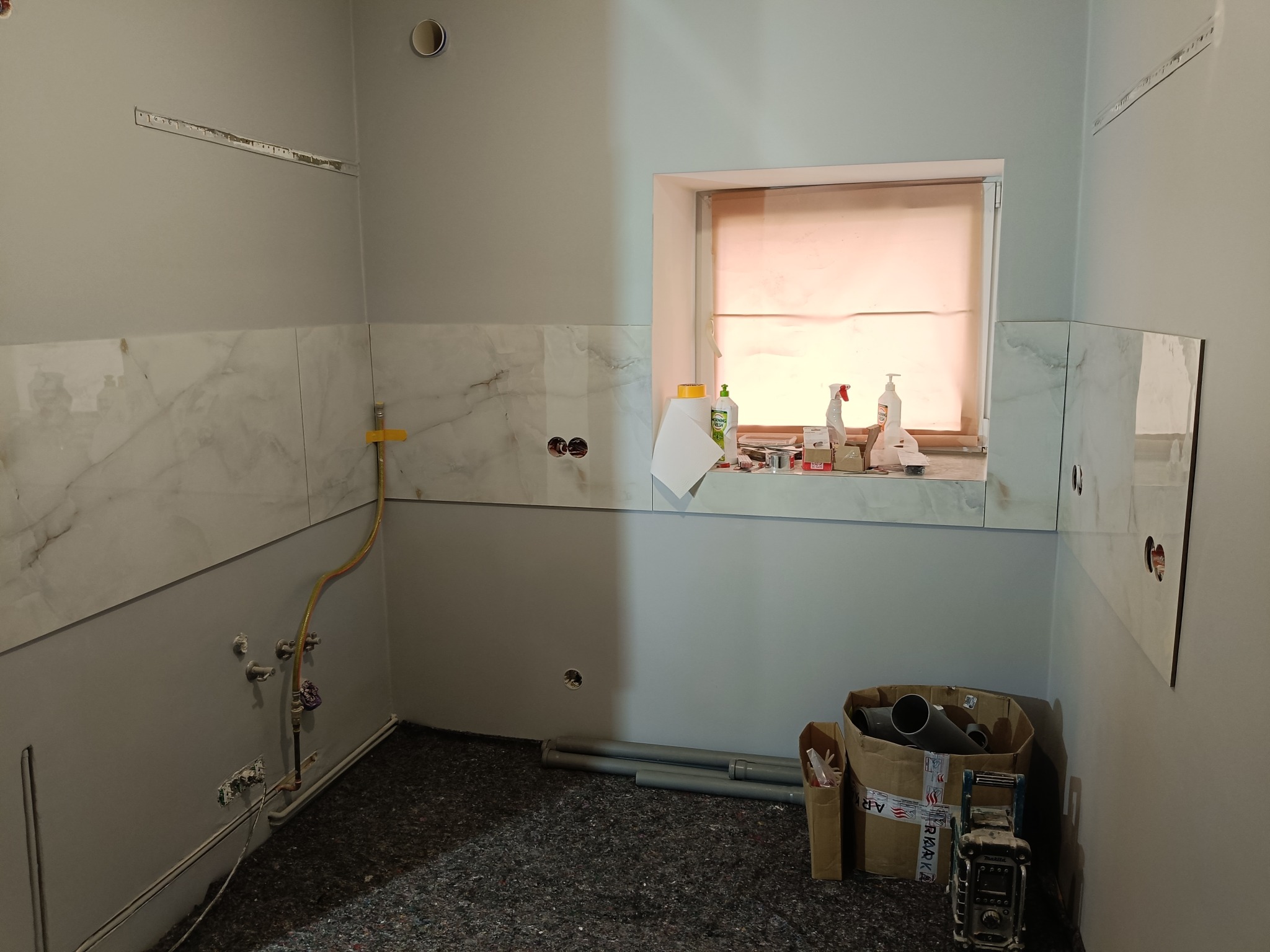 Wnętrze łazienki w trakcie remontu: częściowo położone płytki imitujące marmur, widoczne instalacje hydrauliczne, okno z roletą i narzędzia w kartonach na podłodze.