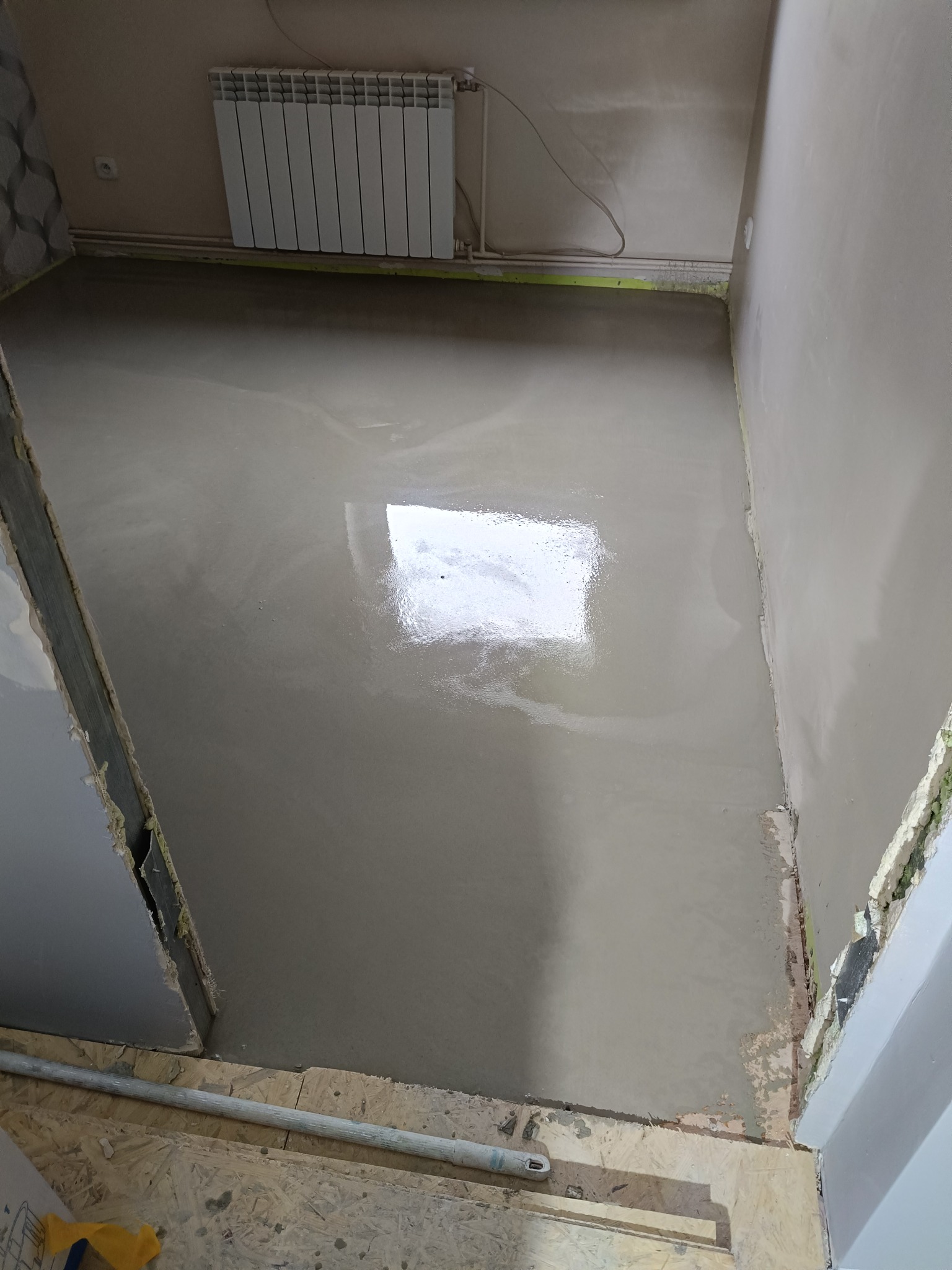 Świeżo wylana, mokra wylewka podłogowa w pomieszczeniu z grzejnikiem. Widoczne przygotowane ściany i podłoże. Ujęcie z góry.