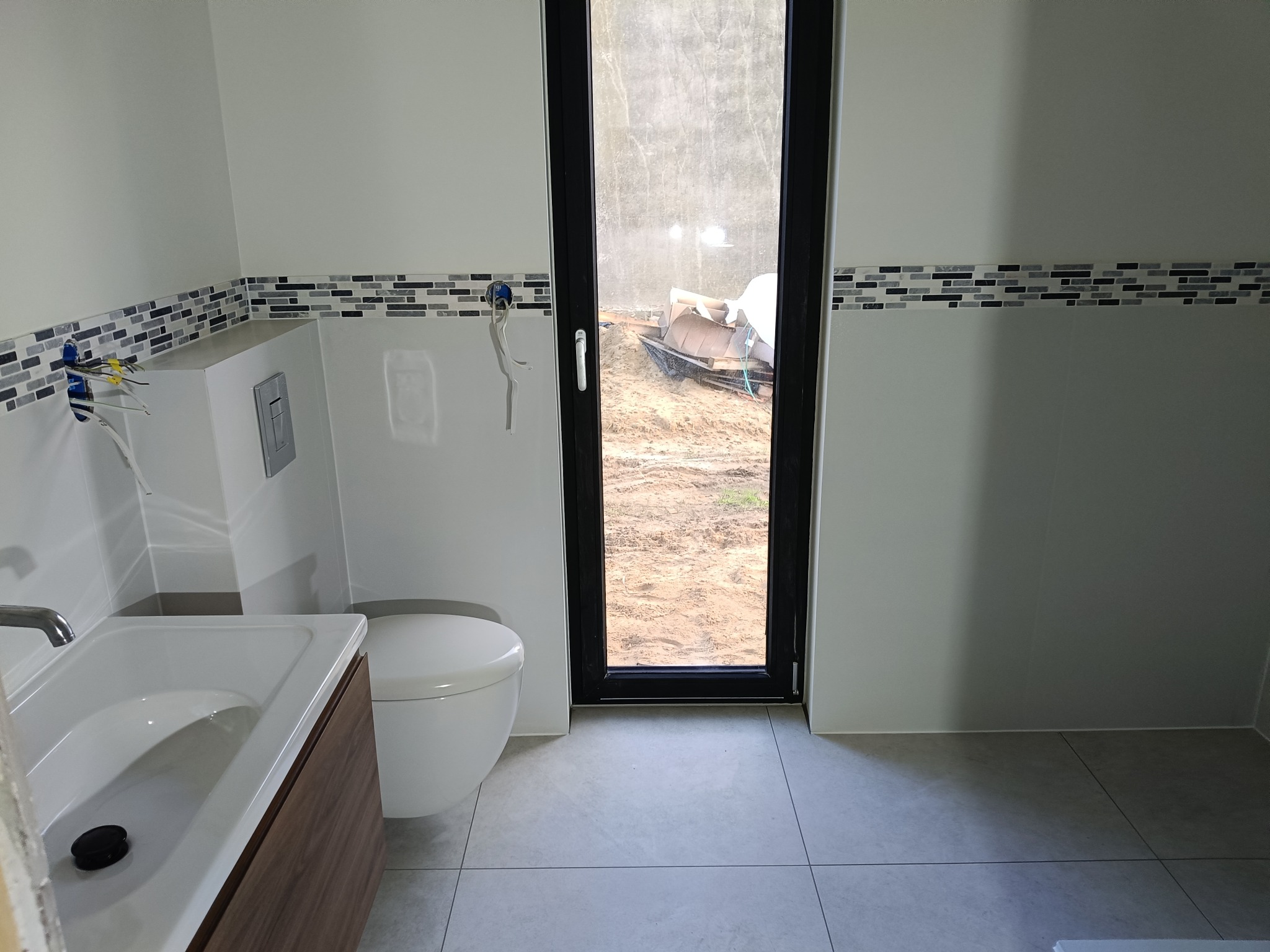 Nowoczesna łazienka z umywalką, toaletą i mozaiką. Widoczne przyłącza hydrauliczne i okno z widokiem na plac budowy. Minimalistyczny design, szare płytki podłogowe.