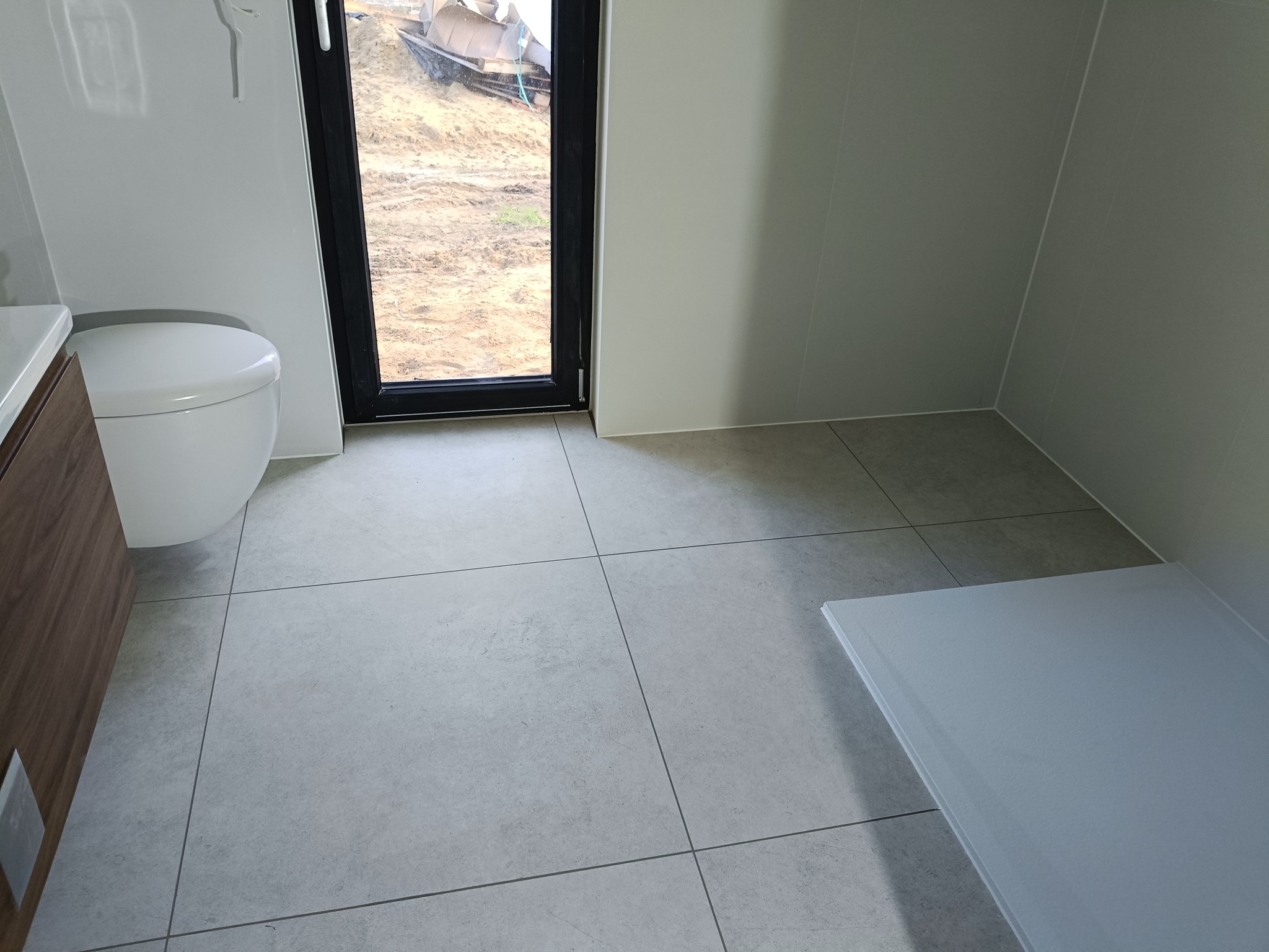 Nowoczesna łazienka z szarą płytką podłogową i kabiną prysznicową. Widoczne okno z widokiem na plac budowy. Minimalistyczny design, jasne ściany.