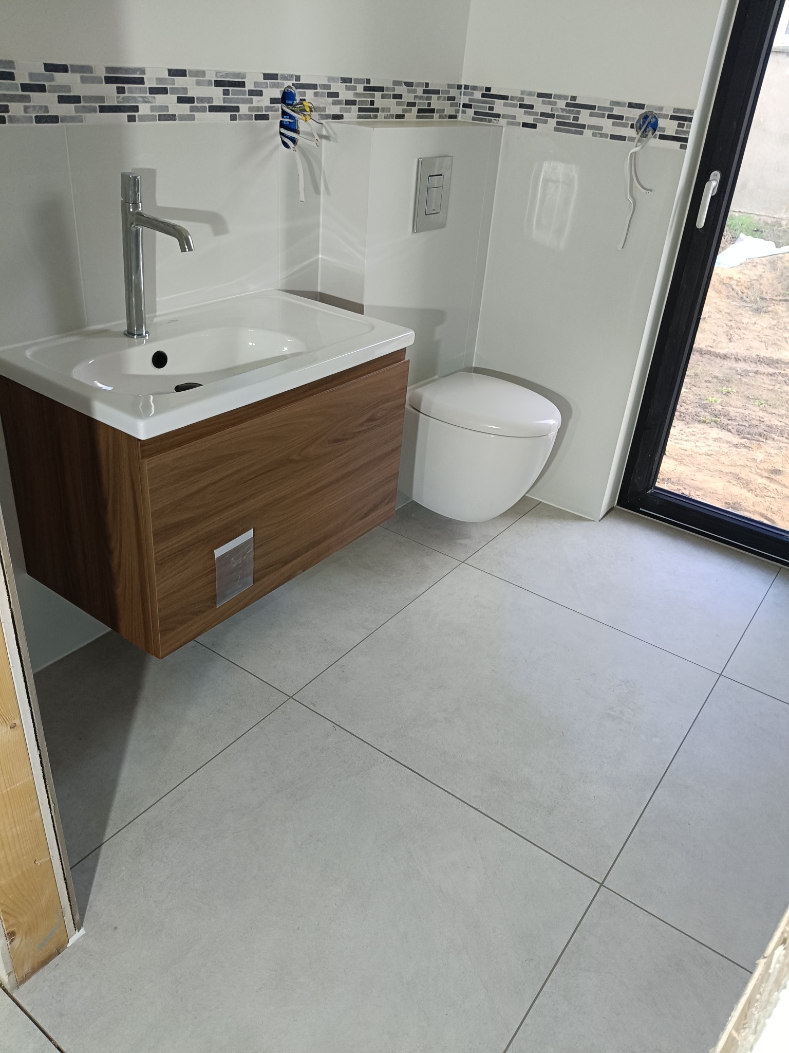 Nowoczesna łazienka z podwieszaną toaletą i umywalką z szafką w kolorze drewna. Na ścianie dekoracyjny pas mozaiki. Jasne płytki podłogowe i duże okno.