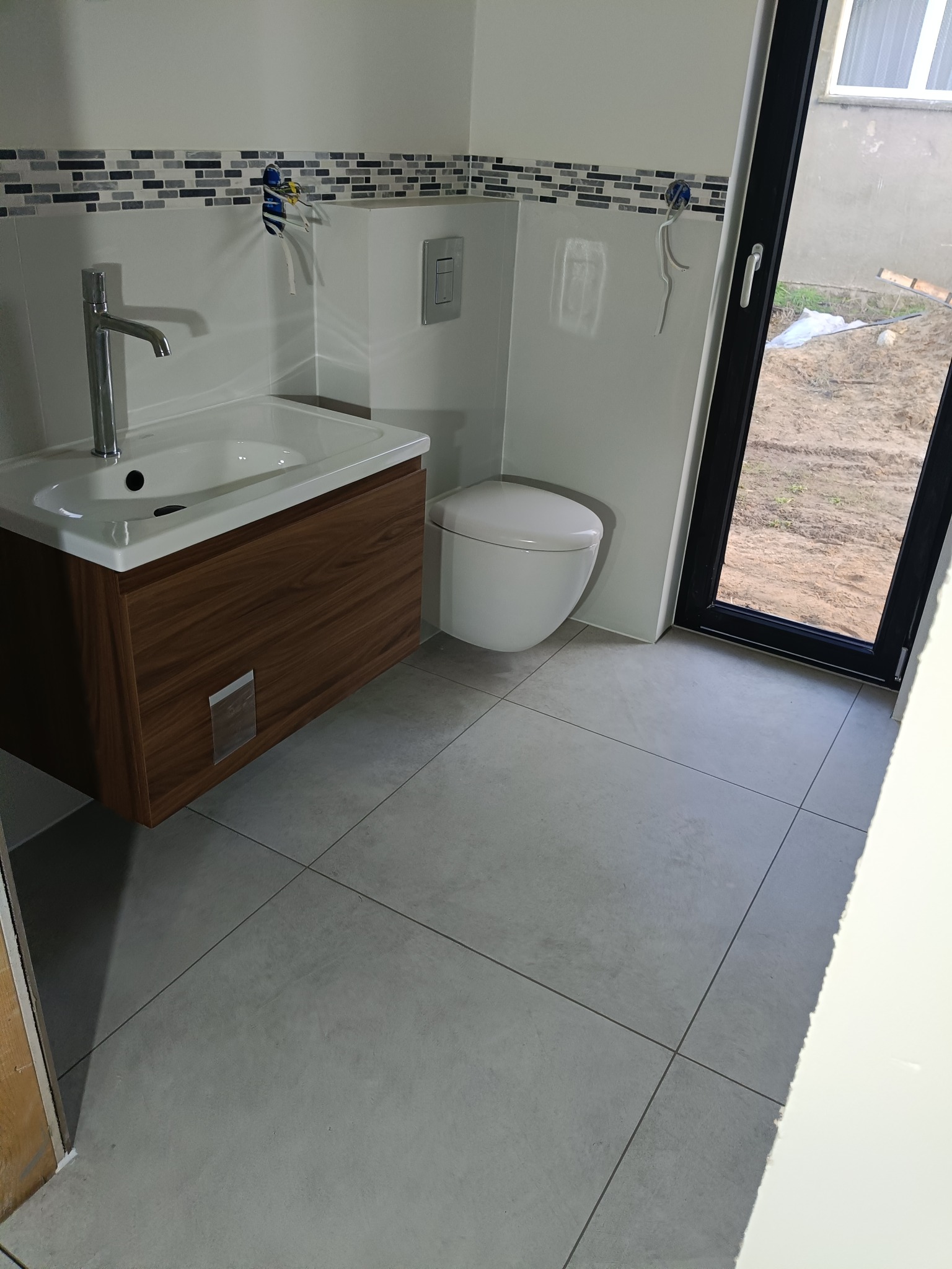 Nowoczesna łazienka po remoncie: podwieszana toaleta, umywalka z szafką z drewna, szara mozaika nad umywalką, szare płytki podłogowe i czarna rama okienna.