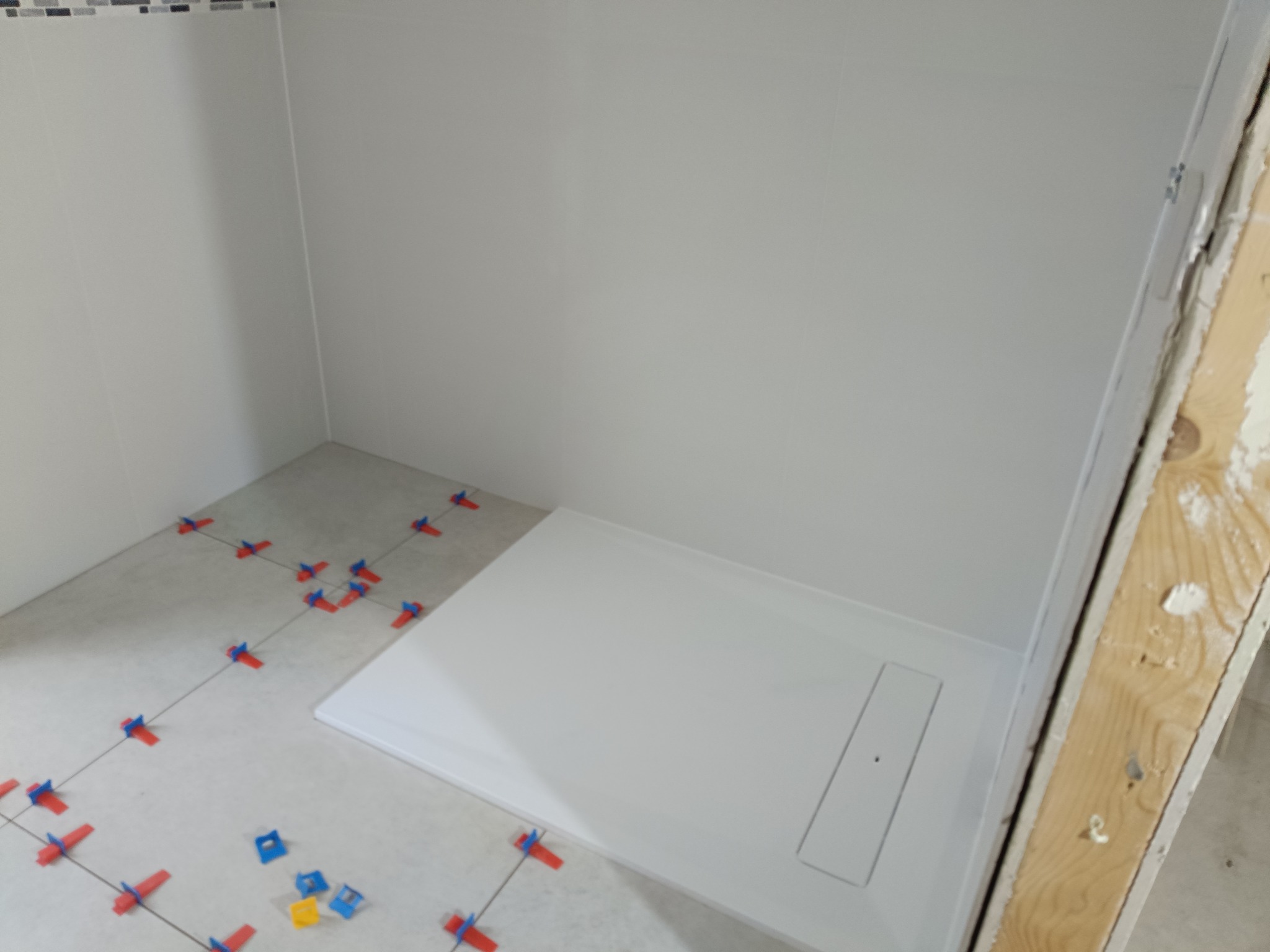 Biały brodzik w narożnej kabinie prysznicowej, płytki podłogowe z systemem poziomowania. Fragment drewnianej ramy drzwi po prawej stronie kadru.