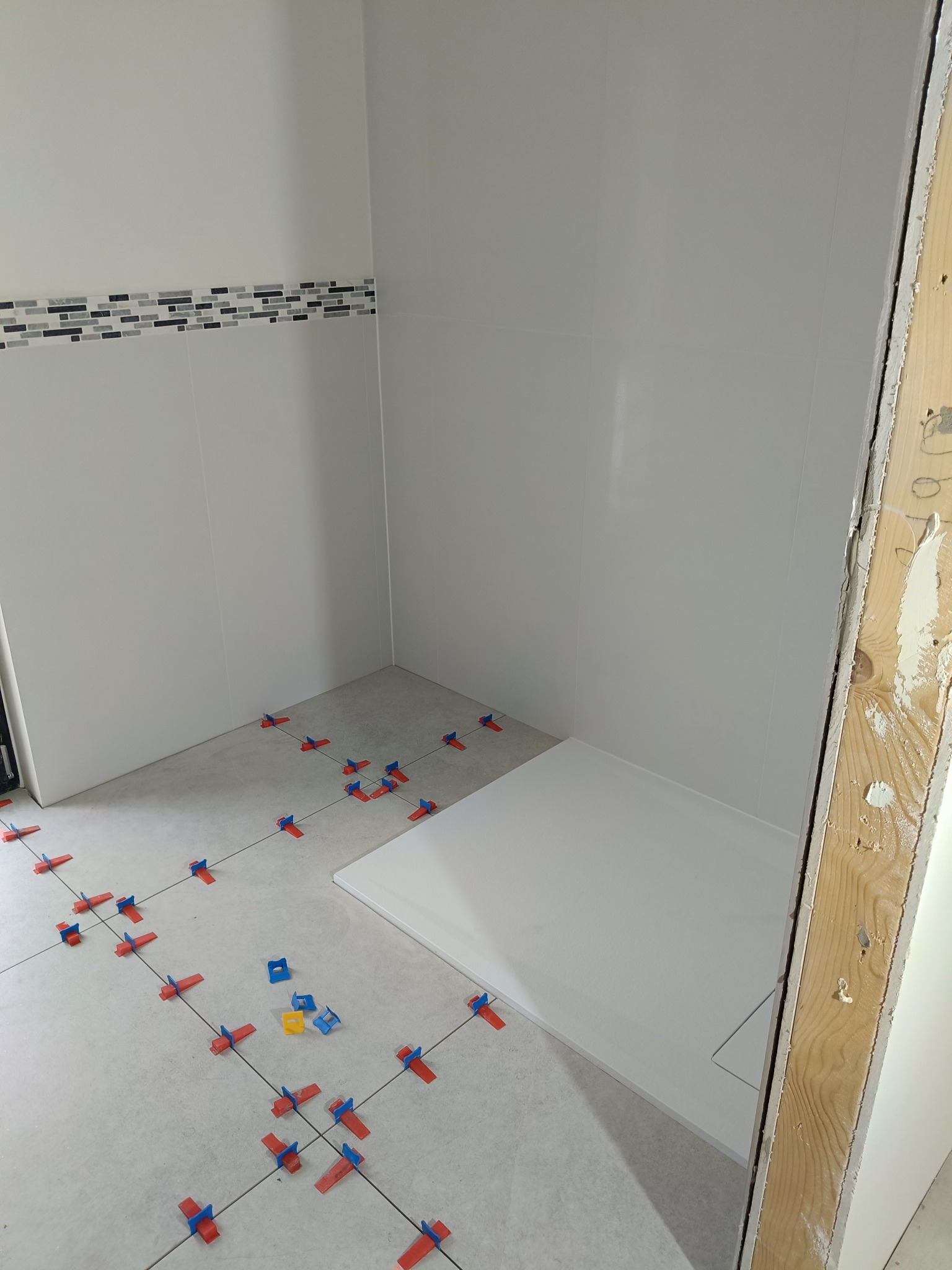 Biała łazienka w trakcie wykańczania: ułożone płytki podłogowe z systemem poziomowania, biały brodzik i pas mozaiki na ścianie. Widoczny fragment drewnianej futryny.