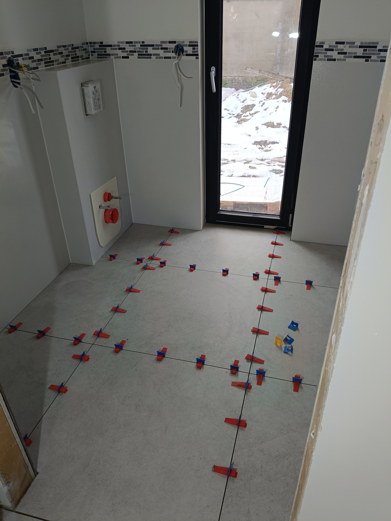 Jasna łazienka w trakcie układania szarych płytek podłogowych z użyciem systemu poziomowania. Widoczne krzyżyki i sznurki wyznaczające linie. Za oknem zimowy krajobraz.