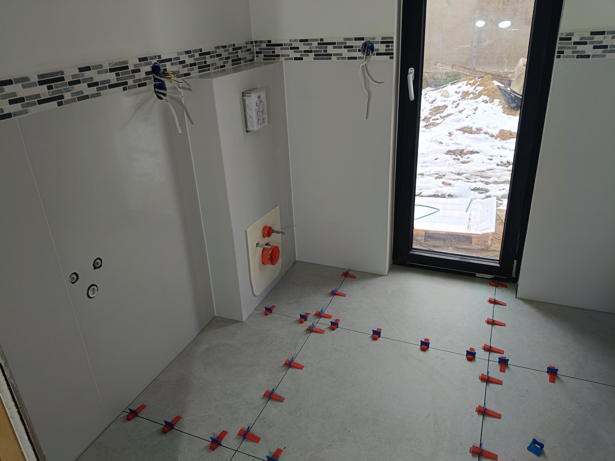 Biała łazienka w trakcie wykańczania: ułożone płytki podłogowe z krzyżykami, częściowo wykończone ściany z mozaiką i instalacją, okno z widokiem na śnieg.