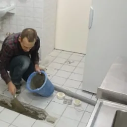 Hydraulik w trakcie naprawy odpływu liniowego w łazience, używając kielni i zaprawy z niebieskiego wiadra. Widoczne płytki i rury.