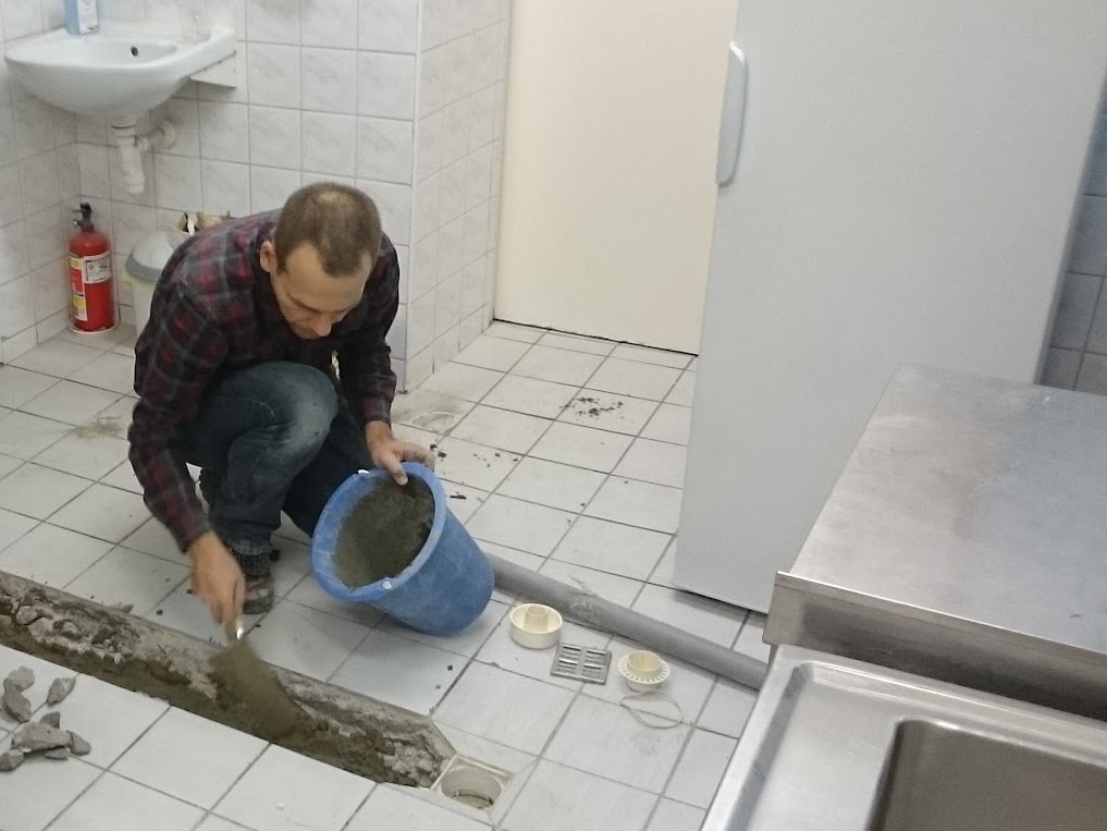 Hydraulik w trakcie naprawy odpływu liniowego w łazience, używając kielni i zaprawy z niebieskiego wiadra. Widoczne płytki i rury.