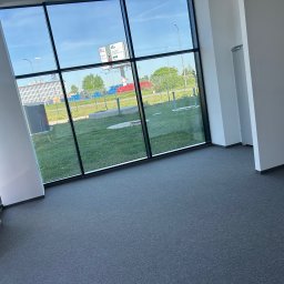 MOTO GASTRO SPÓŁKA Z OGRANICZONĄ ODPOWIEDZIALNOŚCIĄ - Jasne biuro z szarym dywanem i dużym oknem z widokiem na zewnątrz. Minimalistyczny design, nowoczesna przestrzeń biurowa, brak umeblowania, czyste ściany.
