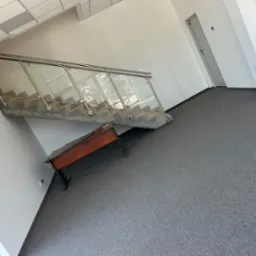 Nowoczesne schody z przeszkloną balustradą w biurze, szara wykładzina podłogowa, białe ściany. Widoczny fragment biurka. Minimalistyczny design, jasne wnętrze.