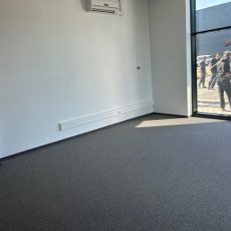 MOTO GASTRO SPÓŁKA Z OGRANICZONĄ ODPOWIEDZIALNOŚCIĄ - Wnętrze biura po remoncie: szara wykładzina, białe ściany z listwami i klimatyzacją. Widok przez szklane drzwi na zewnątrz, gdzie stoją osoby i samochody.