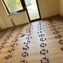 MOTO GASTRO SPÓŁKA Z OGRANICZONĄ ODPOWIEDZIALNOŚCIĄ - Układanie drewnopodobnych płytek na podłodze z użyciem systemu poziomowania. Widoczne niebiesko-czerwone klipsy i kliny. Prace glazurnicze w jasnym wnętrzu z oknem.