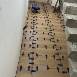 MOTO GASTRO SPÓŁKA Z OGRANICZONĄ ODPOWIEDZIALNOŚCIĄ - Układanie drewnopodobnych płytek na podłodze, z użyciem klipsów poziomujących i poziomicy laserowej. Widoczne schody w tle i kable na podłodze.