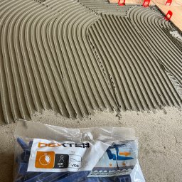 MOTO GASTRO SPÓŁKA Z OGRANICZONĄ ODPOWIEDZIALNOŚCIĄ - Nakładanie kleju grzebieniem na podłogę pod płytki, z użyciem systemu poziomowania płytek (klipsy i kliny). Paczka klipsów Dexter w tle.