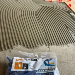 Nakładanie kleju grzebieniem na podłogę pod płytki, z użyciem systemu poziomowania płytek (klipsy i kliny). Paczka klipsów Dexter w tle.