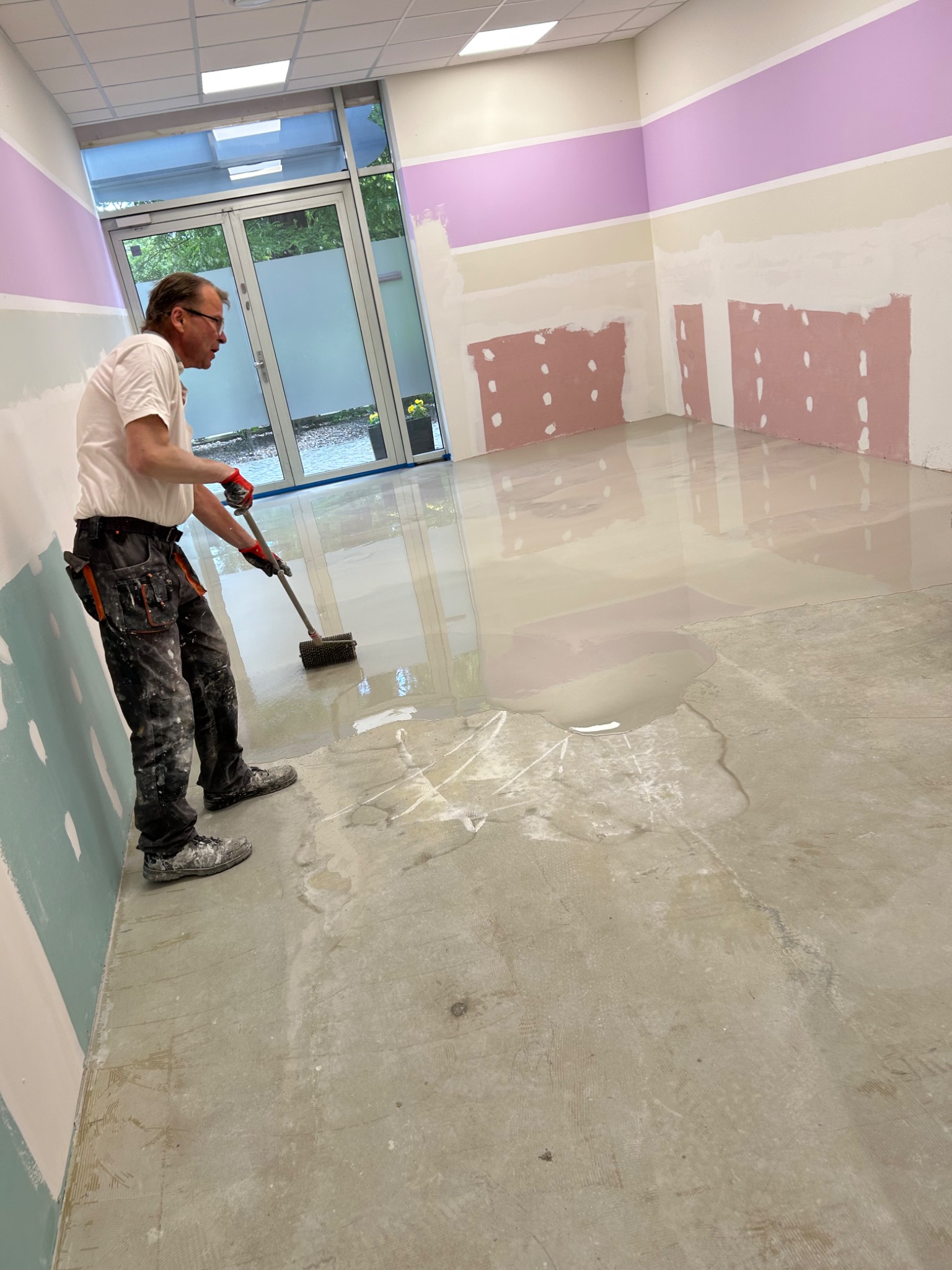 Pracownik rozprowadza wylewkę samopoziomującą na podłodze w pomieszczeniu z przygotowanymi ścianami do malowania. Widoczne narzędzia i etap prac.