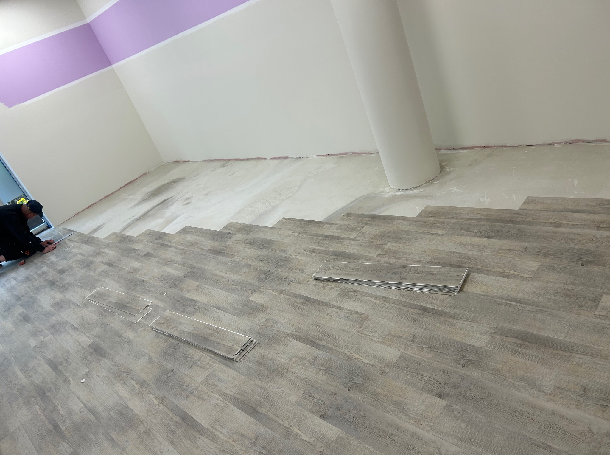 Montaż paneli podłogowych w pomieszczeniu z kolumną. Częściowo ułożona podłoga z szarych paneli imitujących drewno. Widoczny pracownik podczas układania.