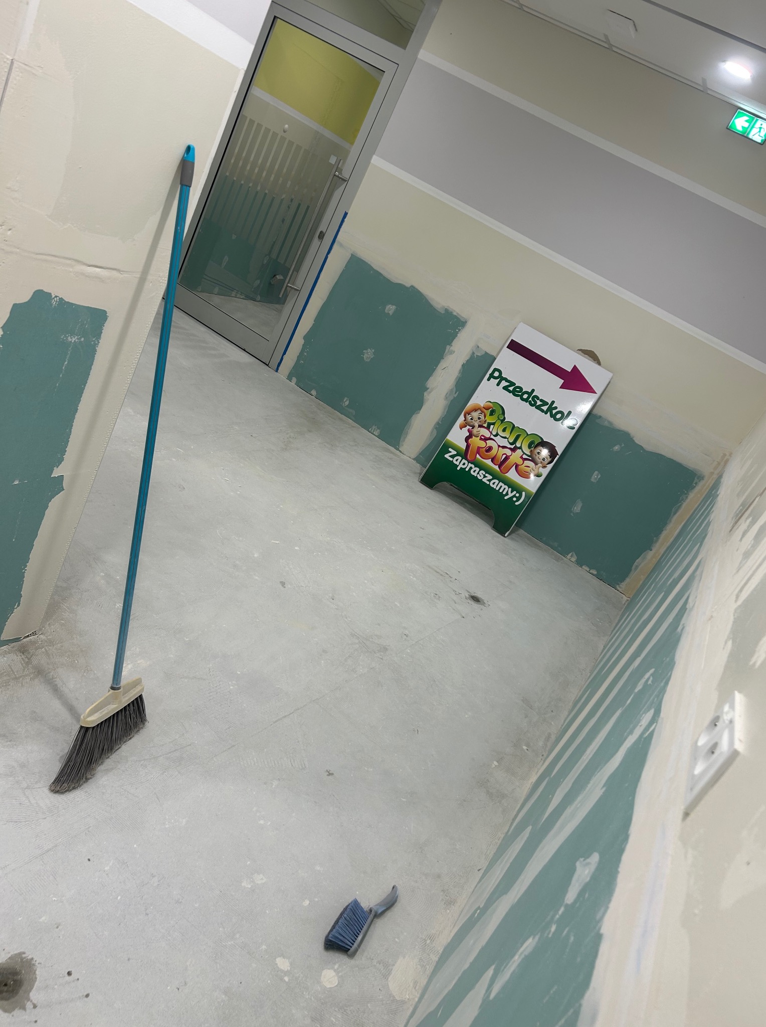 Wnętrze w trakcie wykończenia: ściany z płyt gipsowych pokryte szpachlą, podłoga betonowa, widoczny znak przedszkola i narzędzia. Etap przygotowania do malowania.