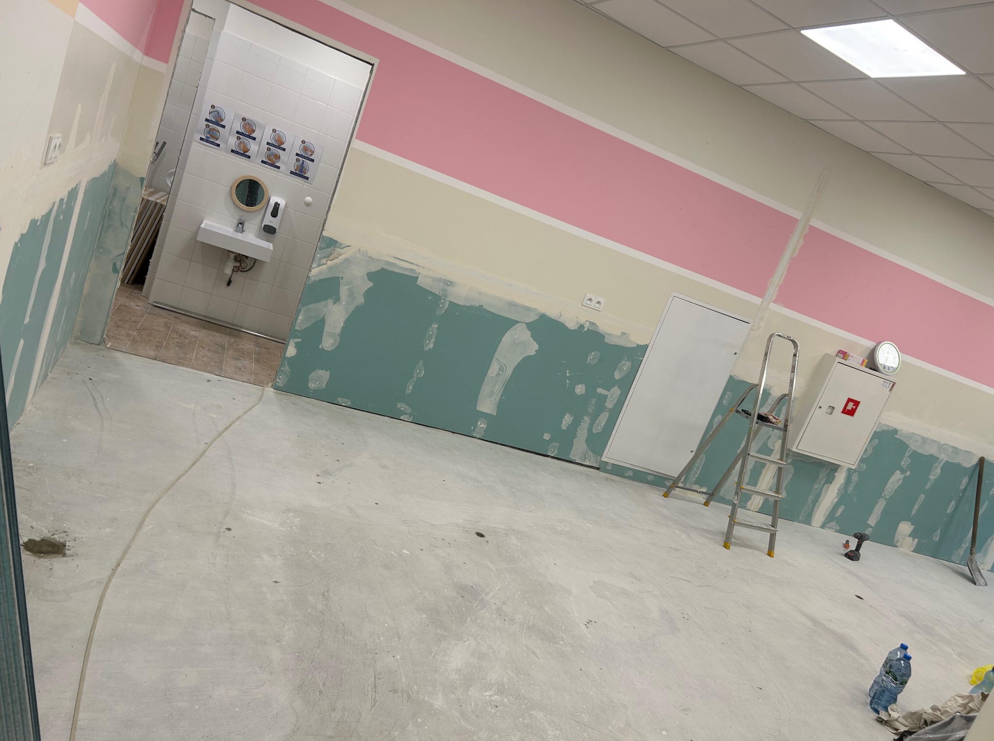 Wnętrze w trakcie remontu z przygotowanymi ścianami do malowania, widoczna drabina, narzędzia i umywalka w tle. Ściany pomalowane w pasy.