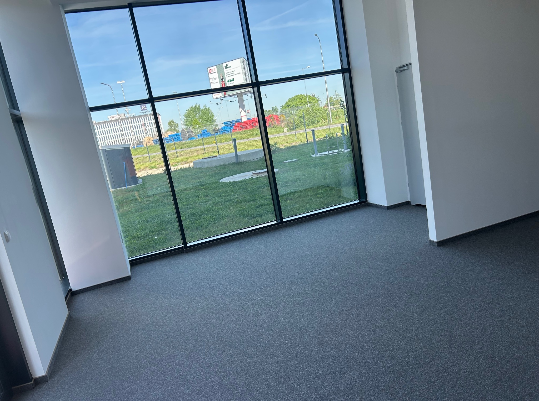 Jasne biuro z szarym dywanem i dużym oknem z widokiem na zewnątrz. Minimalistyczny design, nowoczesna przestrzeń biurowa, brak umeblowania, czyste ściany.