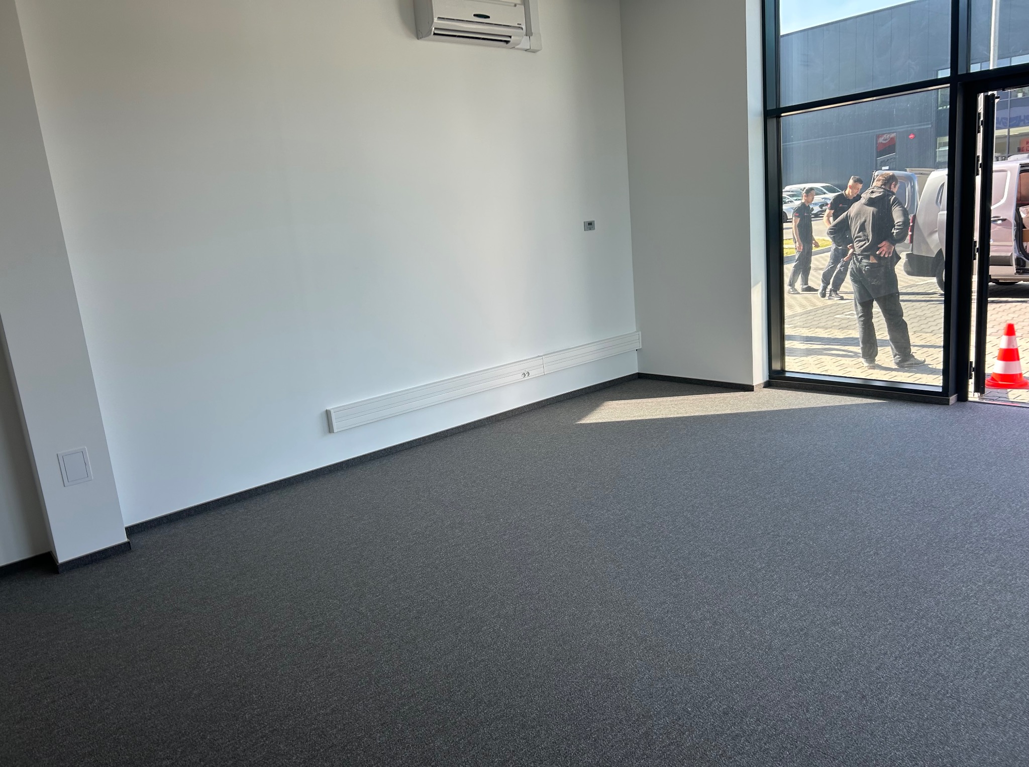 Wnętrze biura po remoncie: szara wykładzina, białe ściany z listwami i klimatyzacją. Widok przez szklane drzwi na zewnątrz, gdzie stoją osoby i samochody.
