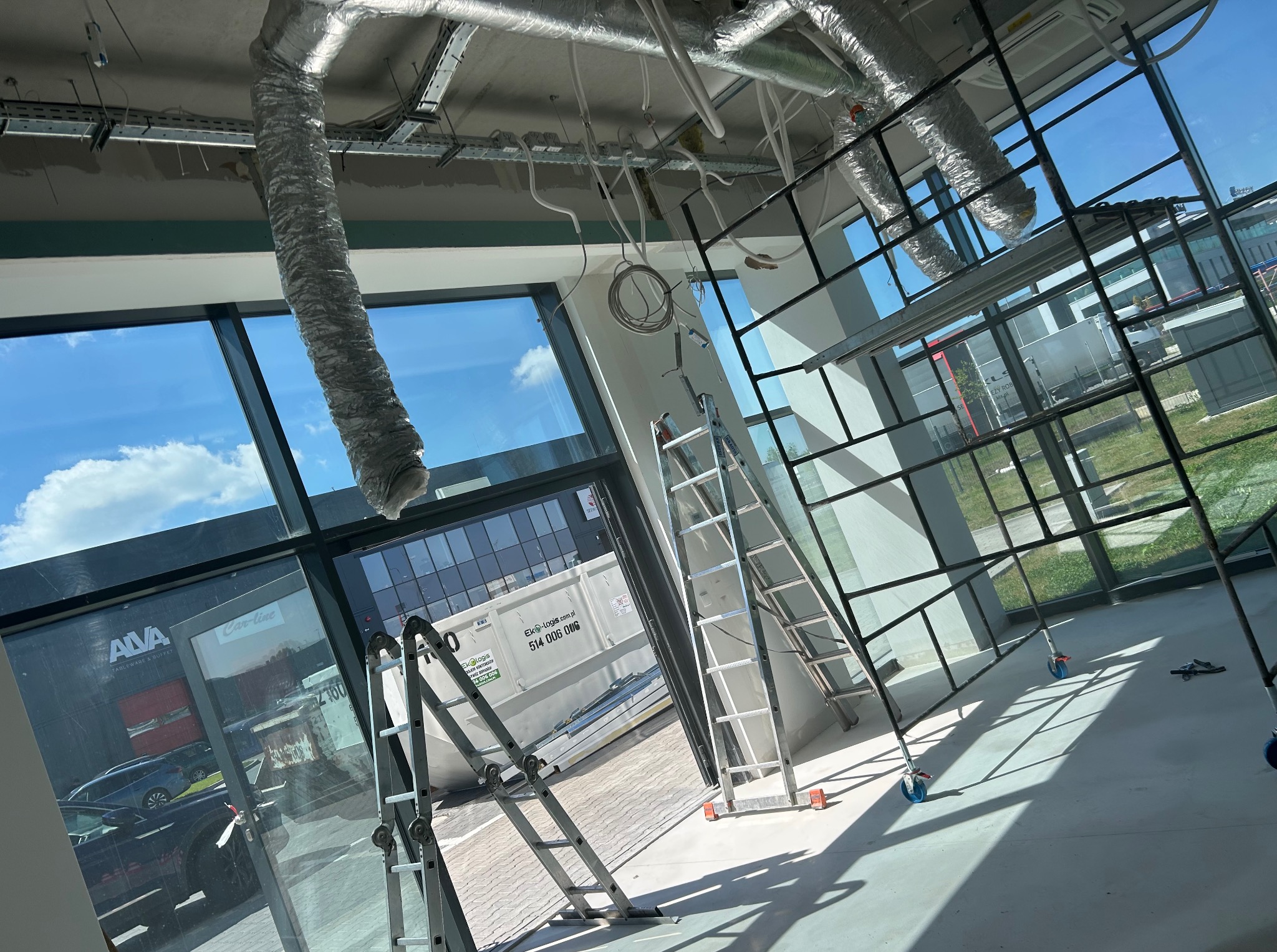 Wnętrze biura w trakcie remontu: widoczne rusztowania, drabiny, instalacje pod sufitem i duże okna z widokiem na zewnątrz. Jasne, przestronne pomieszczenie w trakcie prac wykończeniowych.