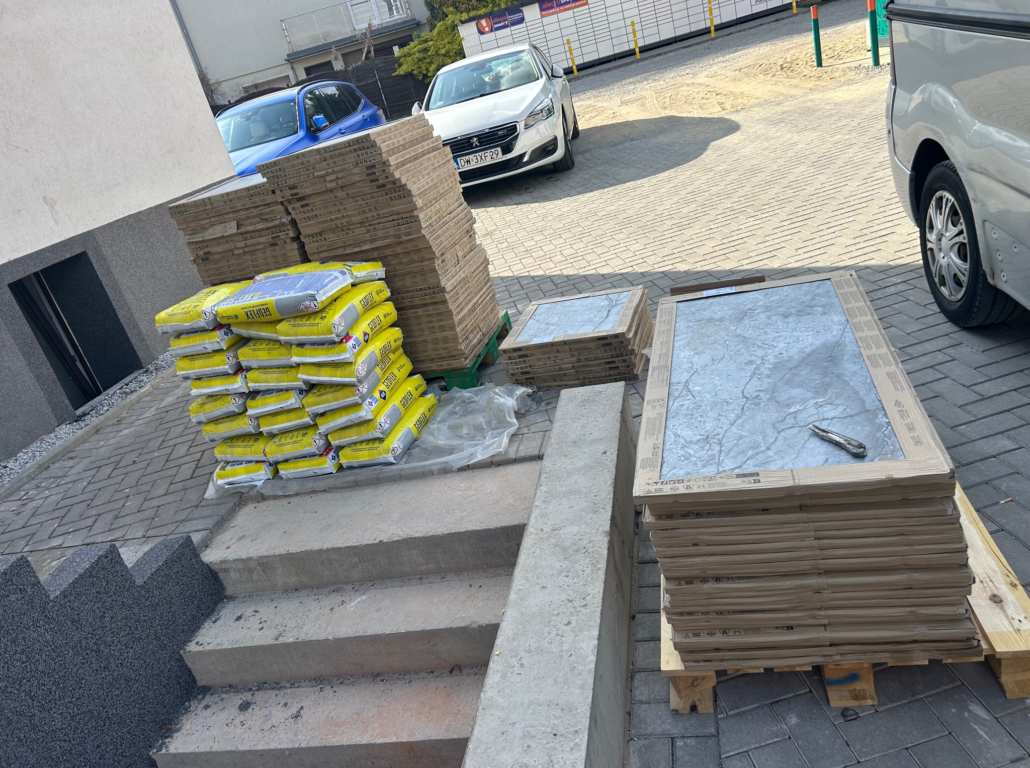Paczki płytek na paletach i worki kleju Geoflex na schodach przed budynkiem. Przygotowanie do prac wykończeniowych na zewnątrz.