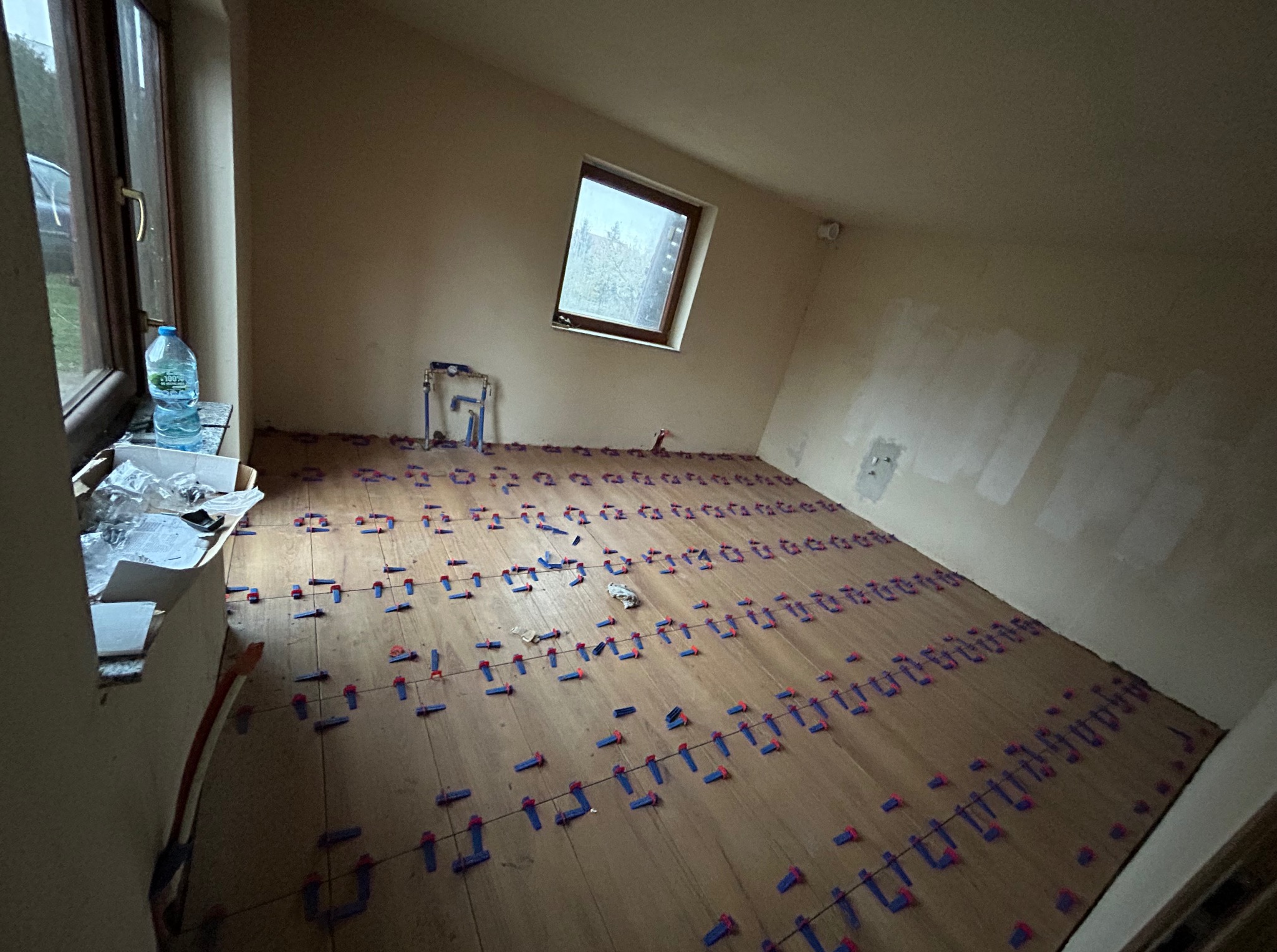 Przygotowanie do układania płytek podłogowych w pokoju na poddaszu, z widocznymi krzyżykami dystansowymi i fragmentem instalacji wodnej przy ścianie.