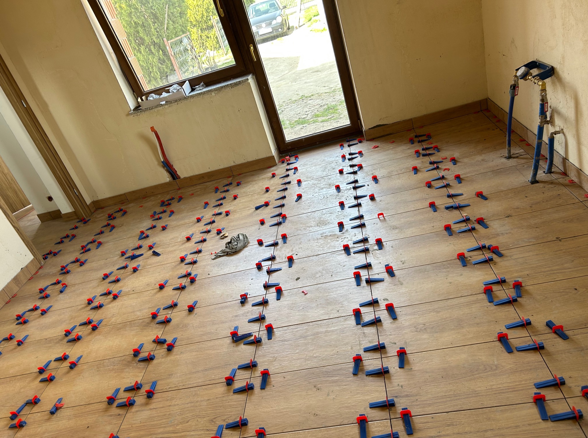 Układanie drewnopodobnych płytek na podłodze z użyciem systemu poziomowania. Widoczne niebiesko-czerwone klipsy i kliny. Prace glazurnicze w jasnym wnętrzu z oknem.