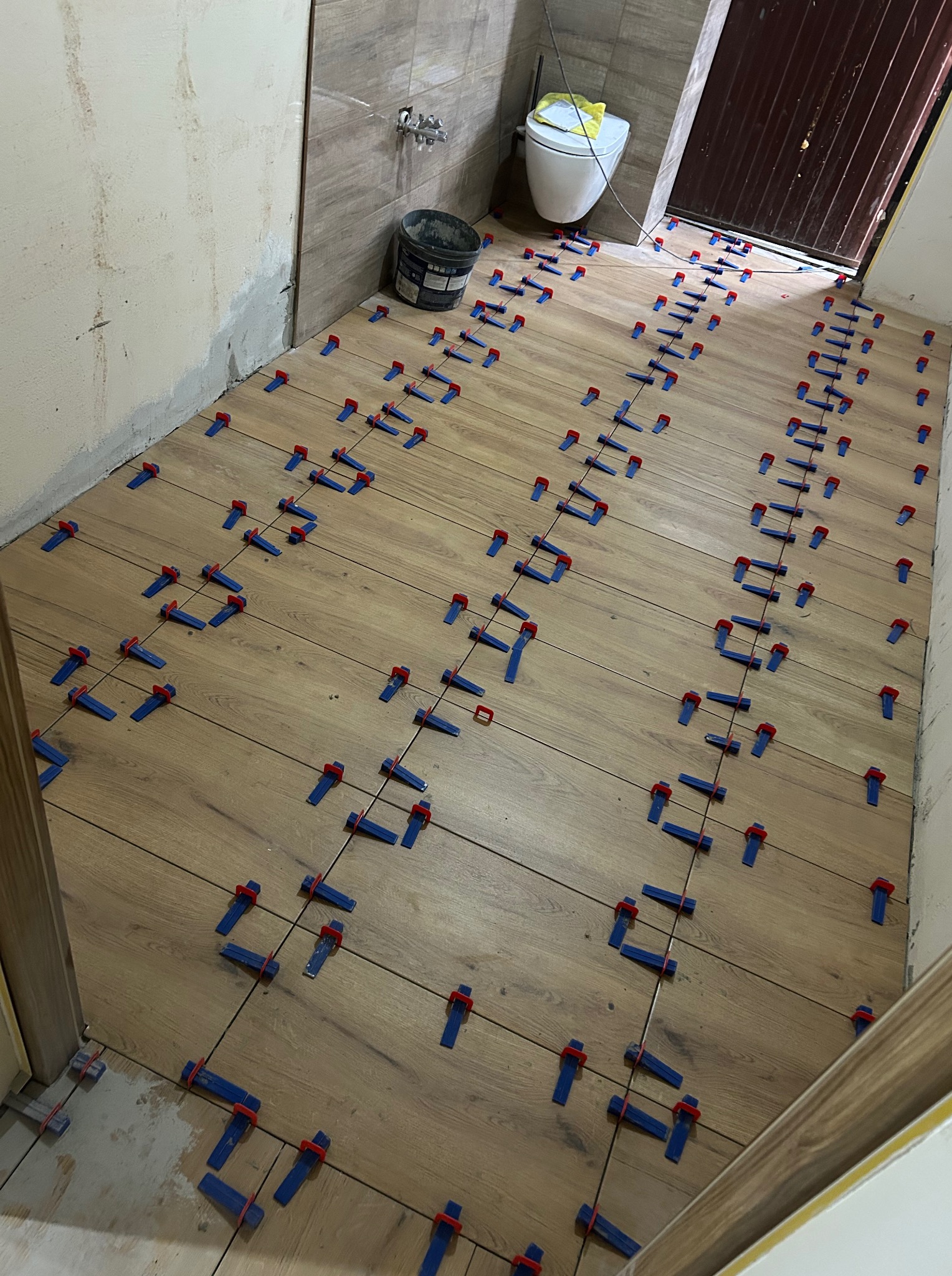 Układanie płytek imitujących drewno na podłodze w łazience z użyciem systemu poziomowania. Widoczne narzędzia glazurnicze i przygotowana ściana pod dalsze prace.