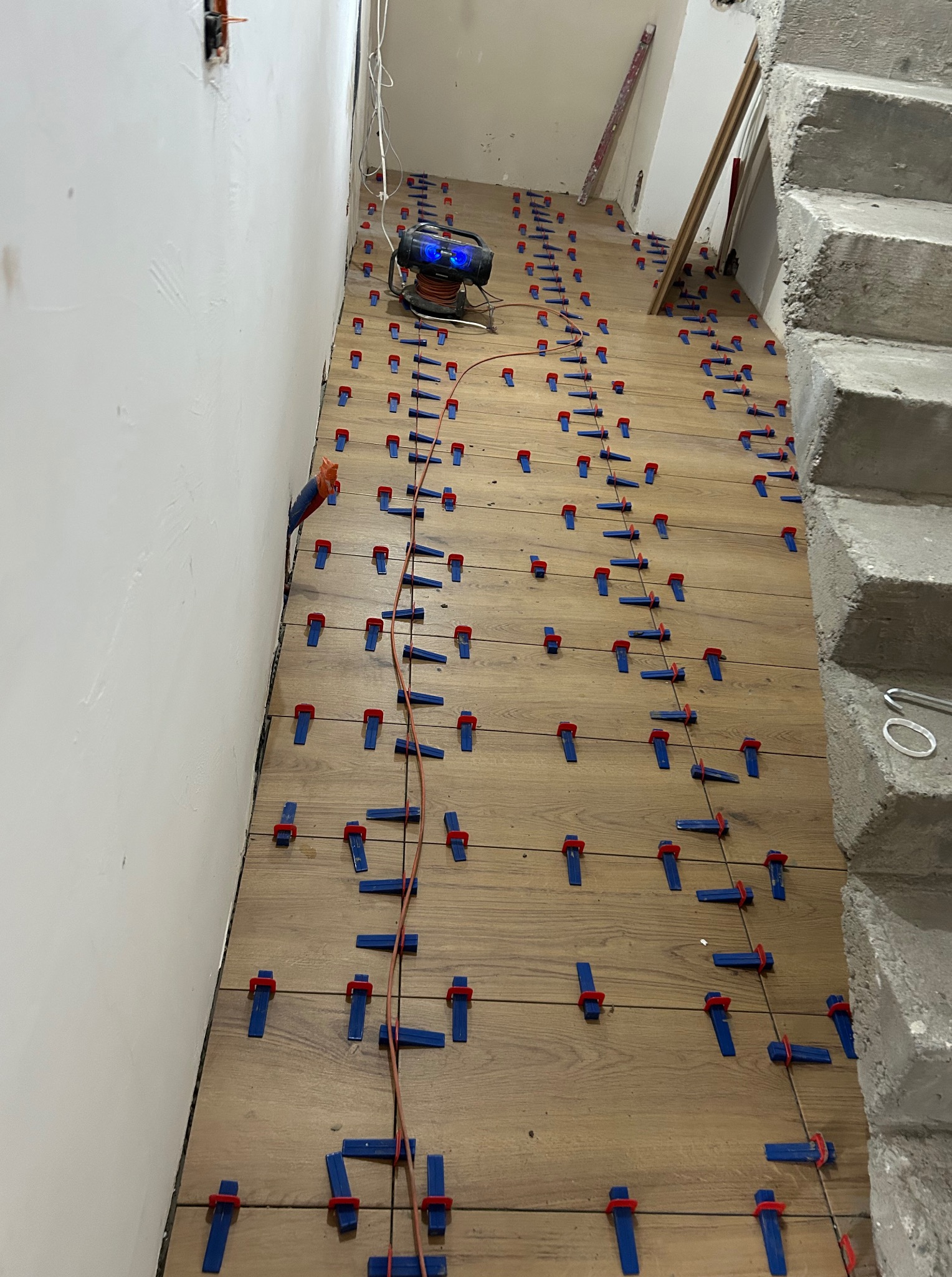Układanie drewnopodobnych płytek na podłodze, z użyciem klipsów poziomujących i poziomicy laserowej. Widoczne schody w tle i kable na podłodze.