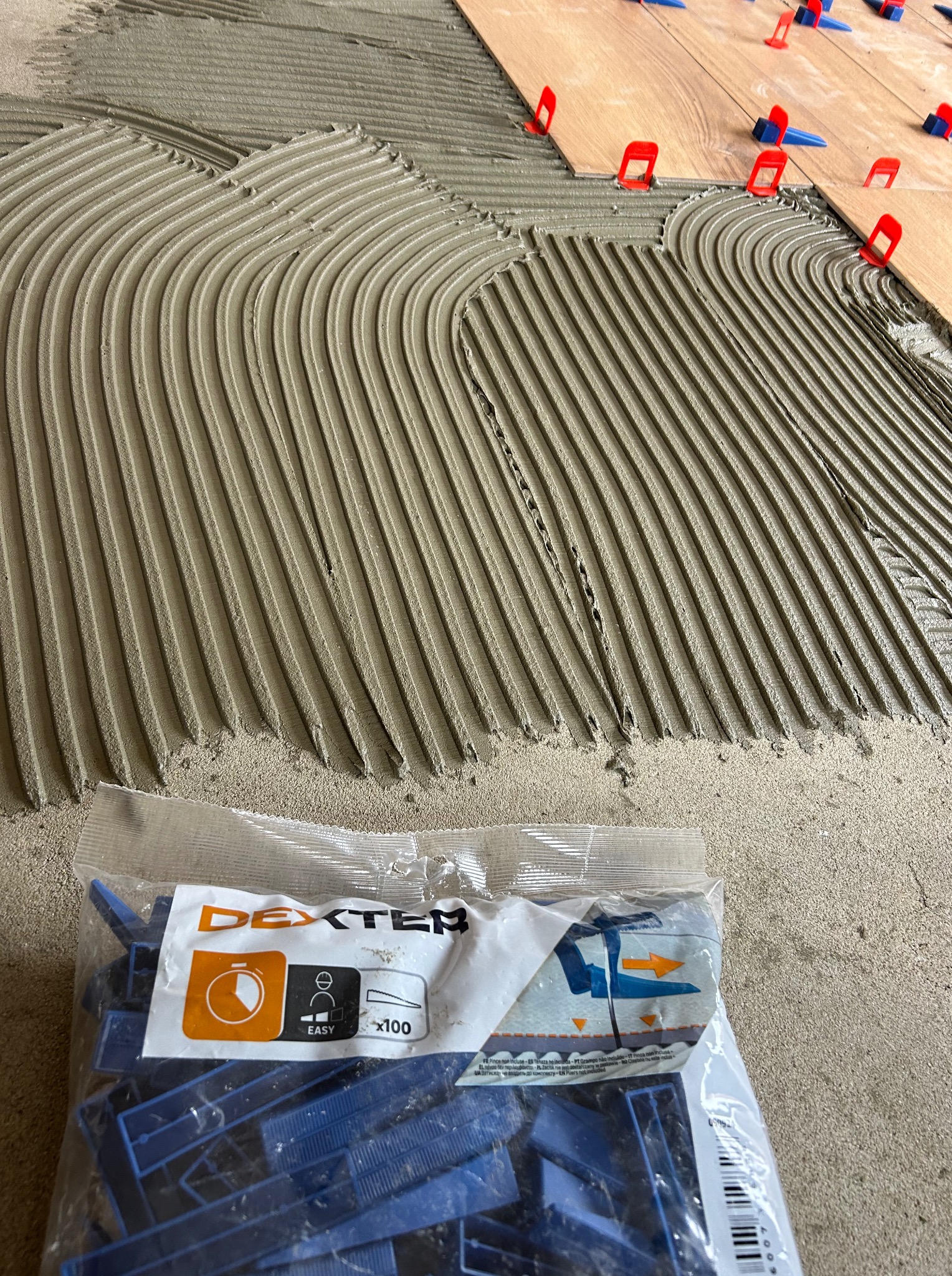 Nakładanie kleju grzebieniem na podłogę pod płytki, z użyciem systemu poziomowania płytek (klipsy i kliny). Paczka klipsów Dexter w tle.