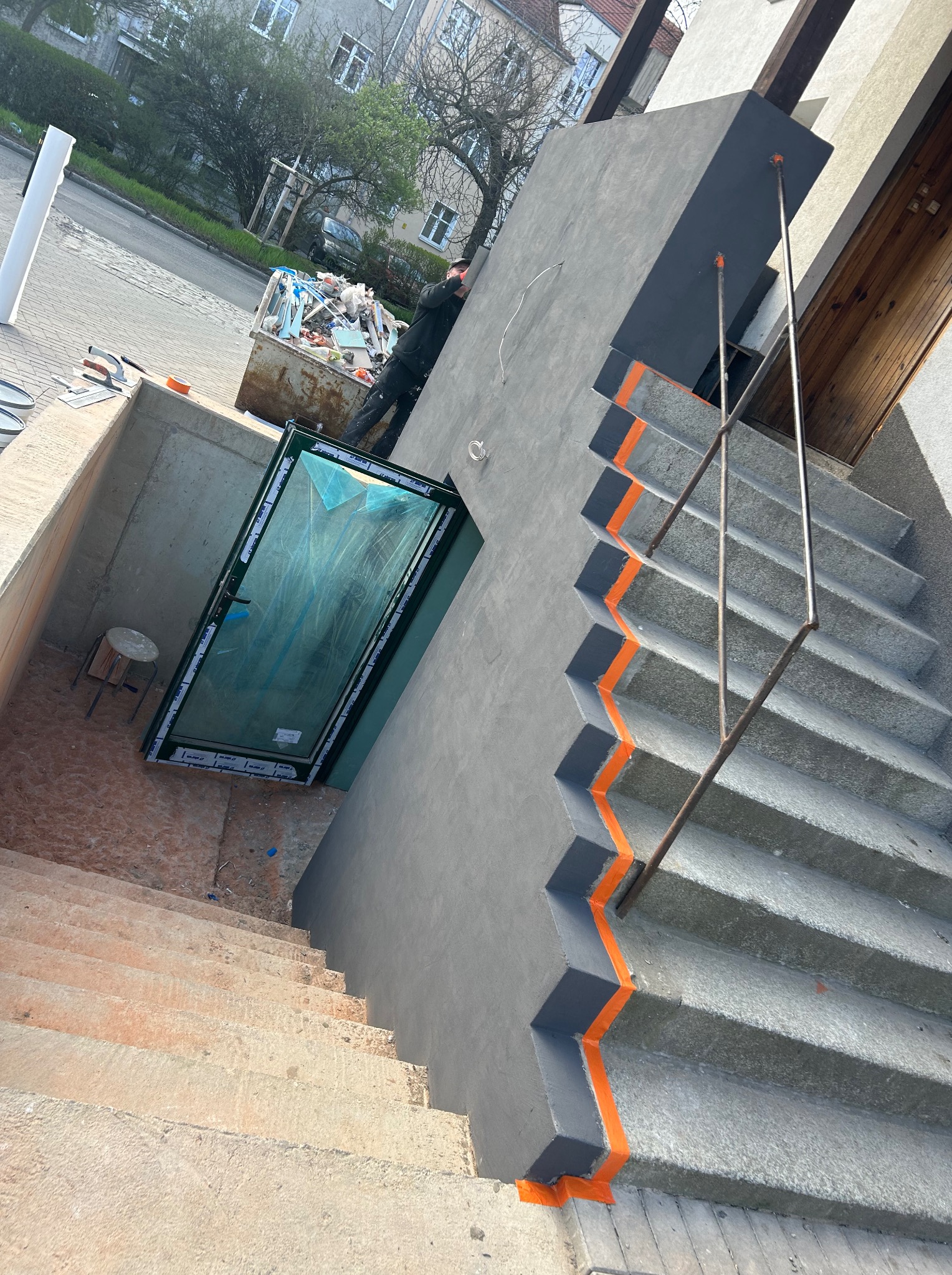 Schody zewnętrzne po remoncie z pomalowanymi elementami na szaro i pomarańczowo, widoczne nowe drzwi do piwnicy i kontener na śmieci.