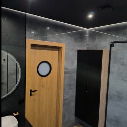 EWK MARCIN NALAZEK - Nowoczesna łazienka z szarymi płytkami, drewnianą podłogą i drzwiami z okrągłym oknem. Czarna szafka wisząca i okrągłe lustro dodają elegancji.