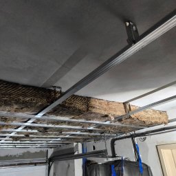 EWK MARCIN NALAZEK - Montaż izolacji z wełny mineralnej na metalowym ruszcie sufitowym w Kałdus. Widoczne przewody elektryczne i fragment ściany. Prace wykończeniowe.