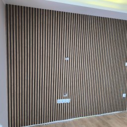 EWK MARCIN NALAZEK - Ściana wykończona panelami lamelowymi w odcieniach drewna, z widocznymi gniazdkami elektrycznymi i fragmentem drzwi po prawej stronie. Nowoczesny design wnętrza.