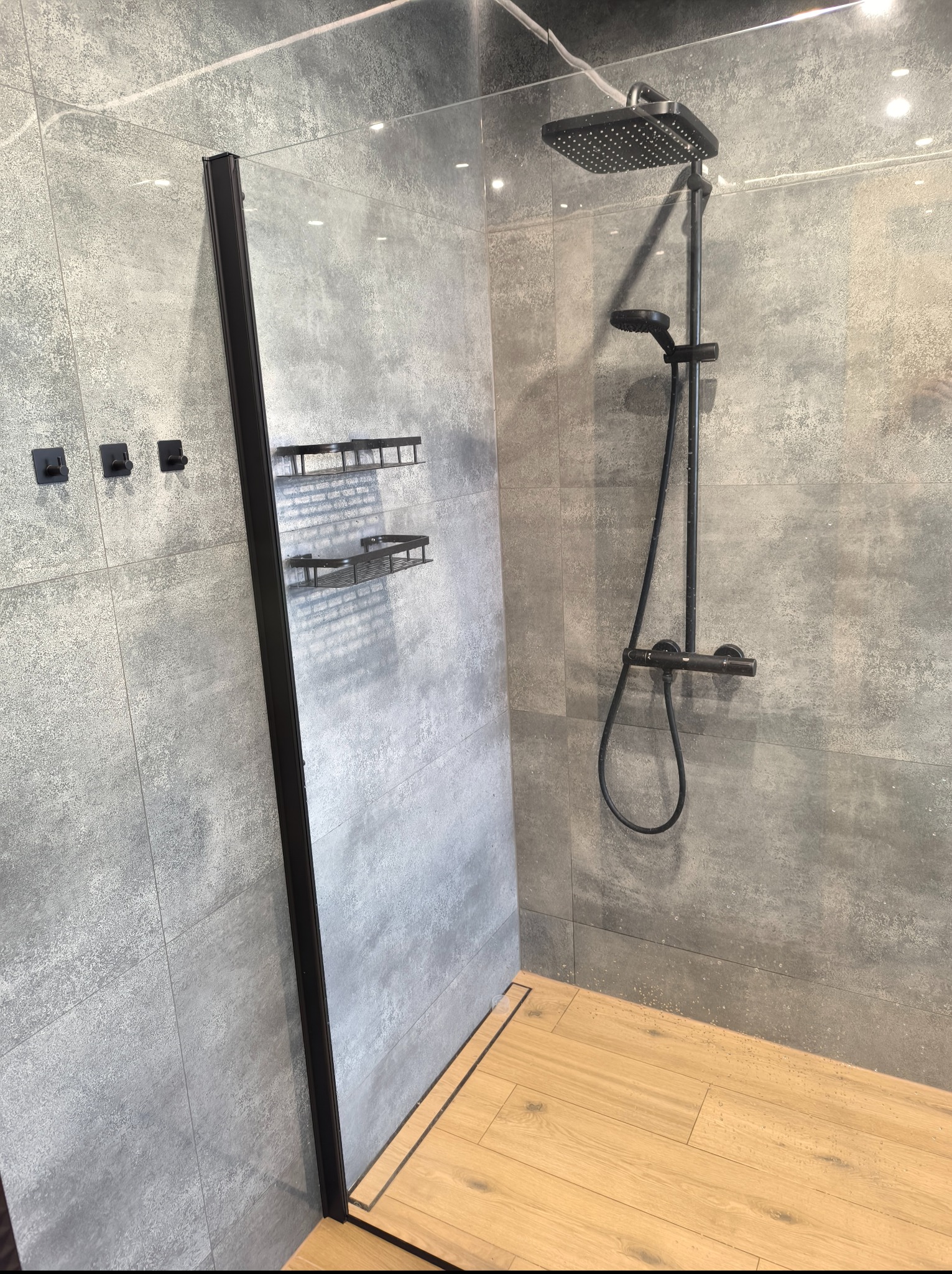 Nowoczesna kabina prysznicowa walk-in z czarną armaturą i szklaną ścianką na tle szarych płytek. Widoczny odpływ liniowy i drewnopodobna podłoga.