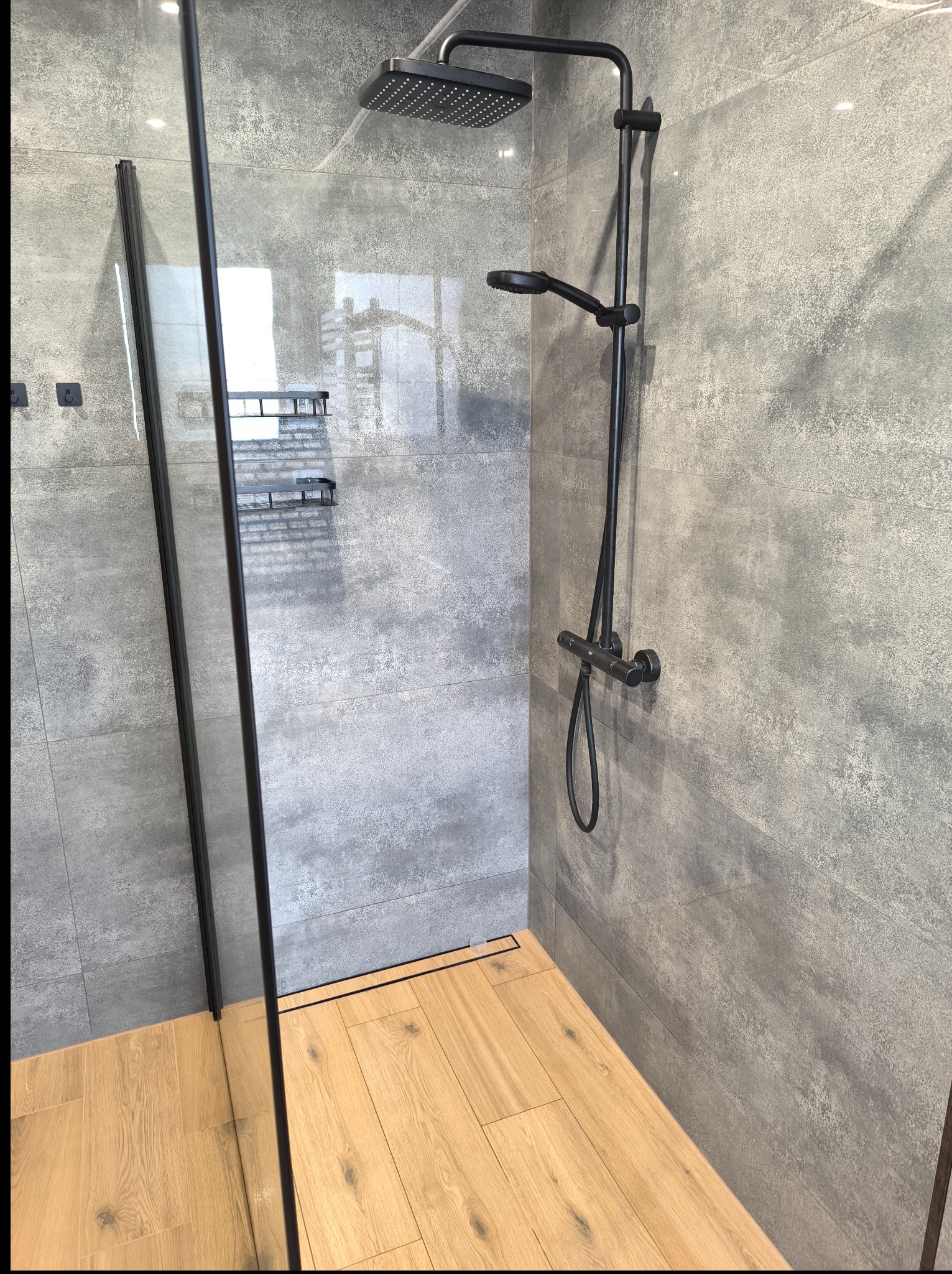 Nowoczesna kabina prysznicowa z szarą, betonową glazurą i drewnopodobną podłogą. Czarna armatura i szklana ściana kabiny. Minimalistyczny design.