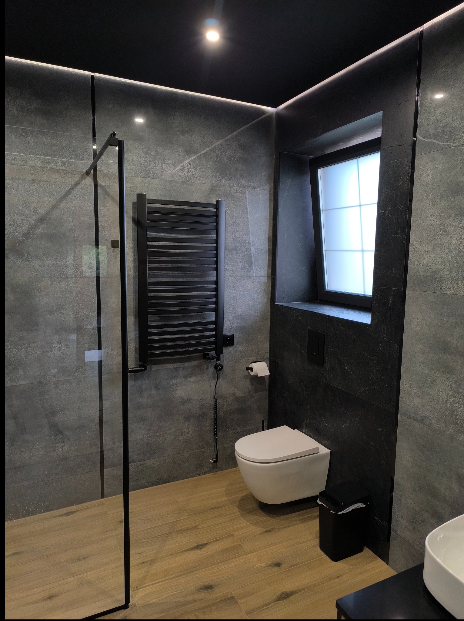 Nowoczesna łazienka z szarą płytką imitującą kamień, drewnopodobną podłogą i czarnymi akcentami: grzejnik, rama okna, kabina prysznicowa. Minimalistyczny design.