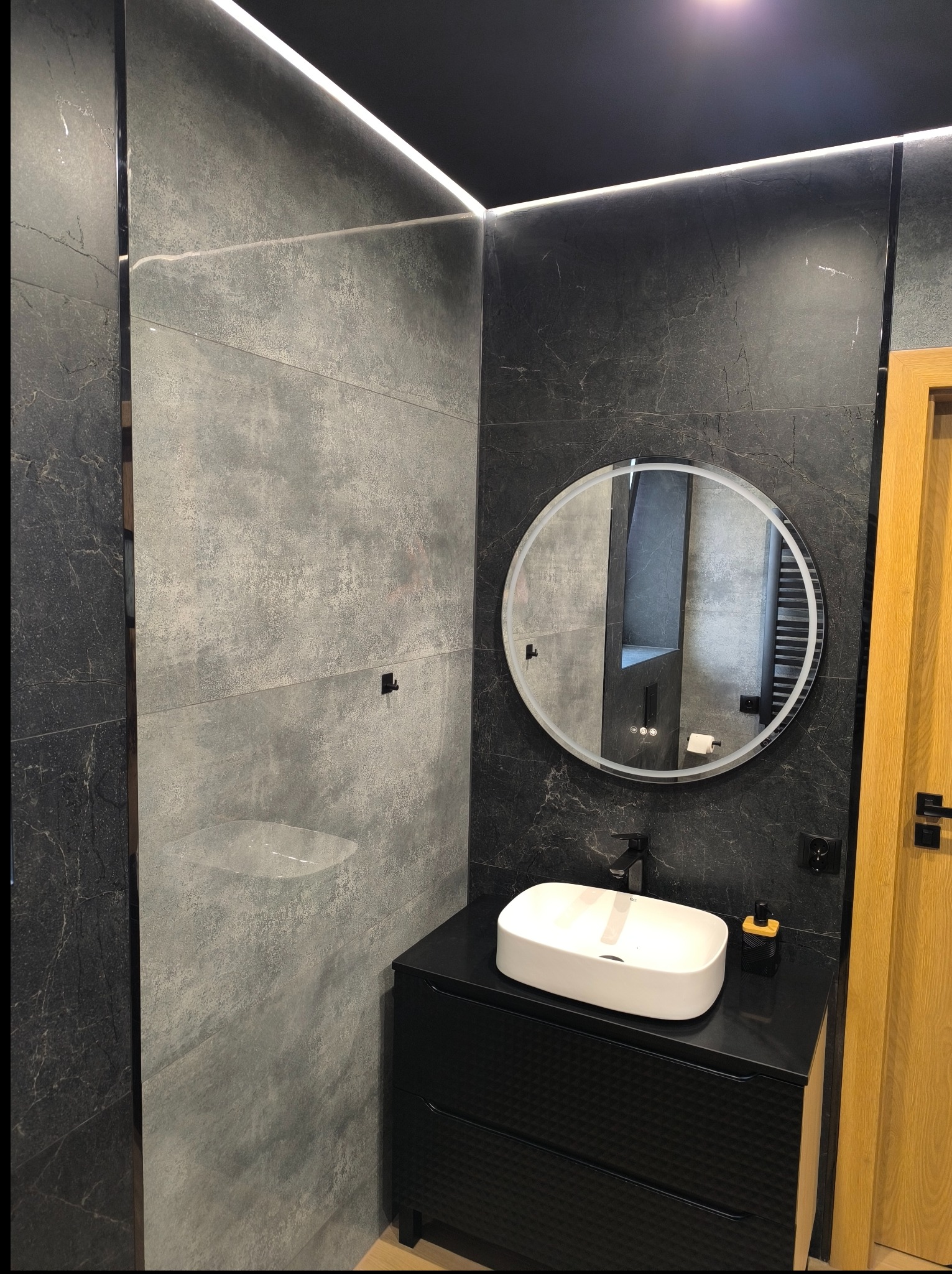 Nowoczesna łazienka z szarymi płytkami imitującymi kamień, okrągłym lustrem z podświetleniem LED i czarną szafką pod umywalką. Minimalistyczny design z akcentami drewna.