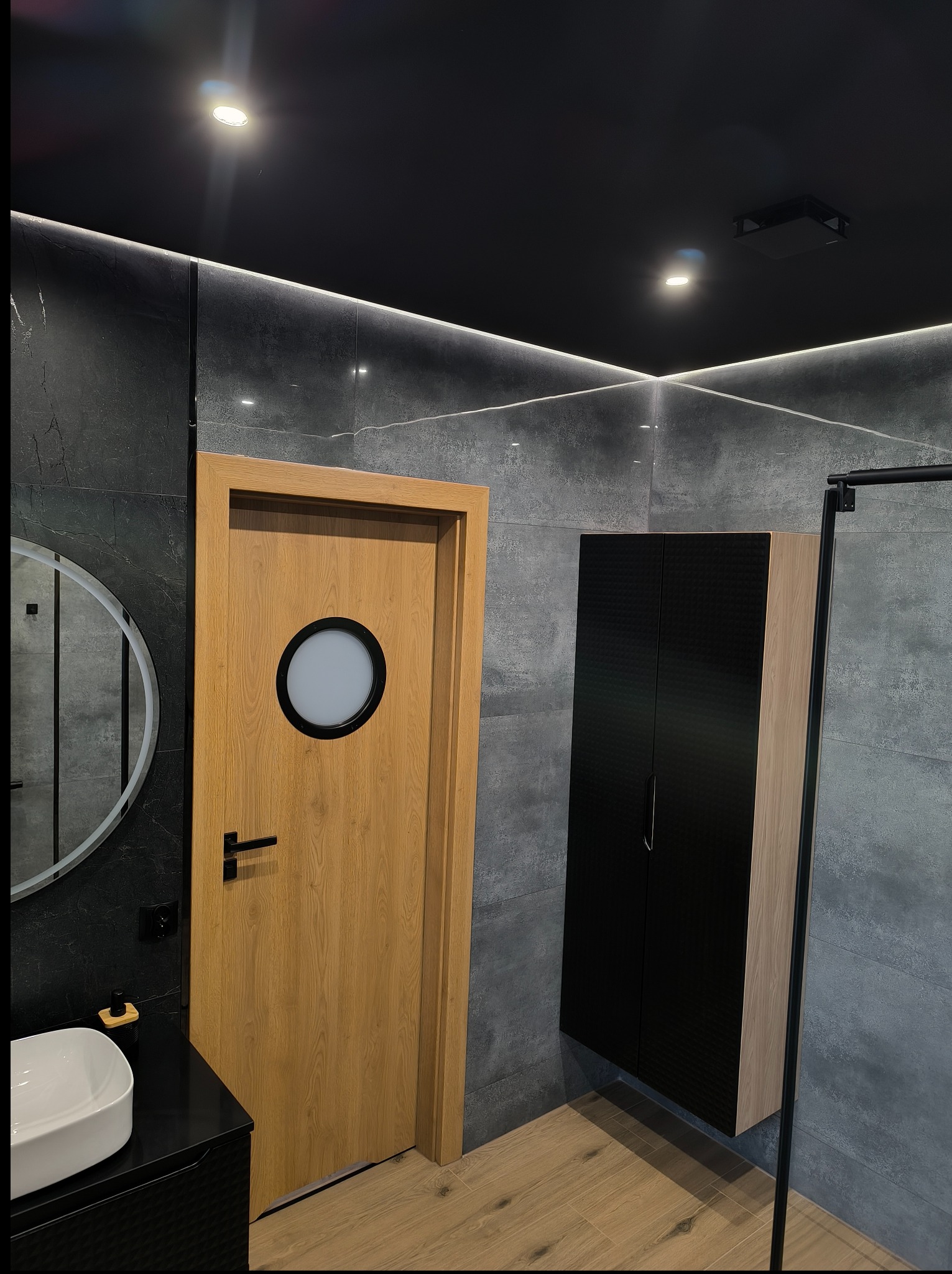 Nowoczesna łazienka z szarymi płytkami, drewnianą podłogą i drzwiami z okrągłym oknem. Czarna szafka wisząca i okrągłe lustro dodają elegancji.