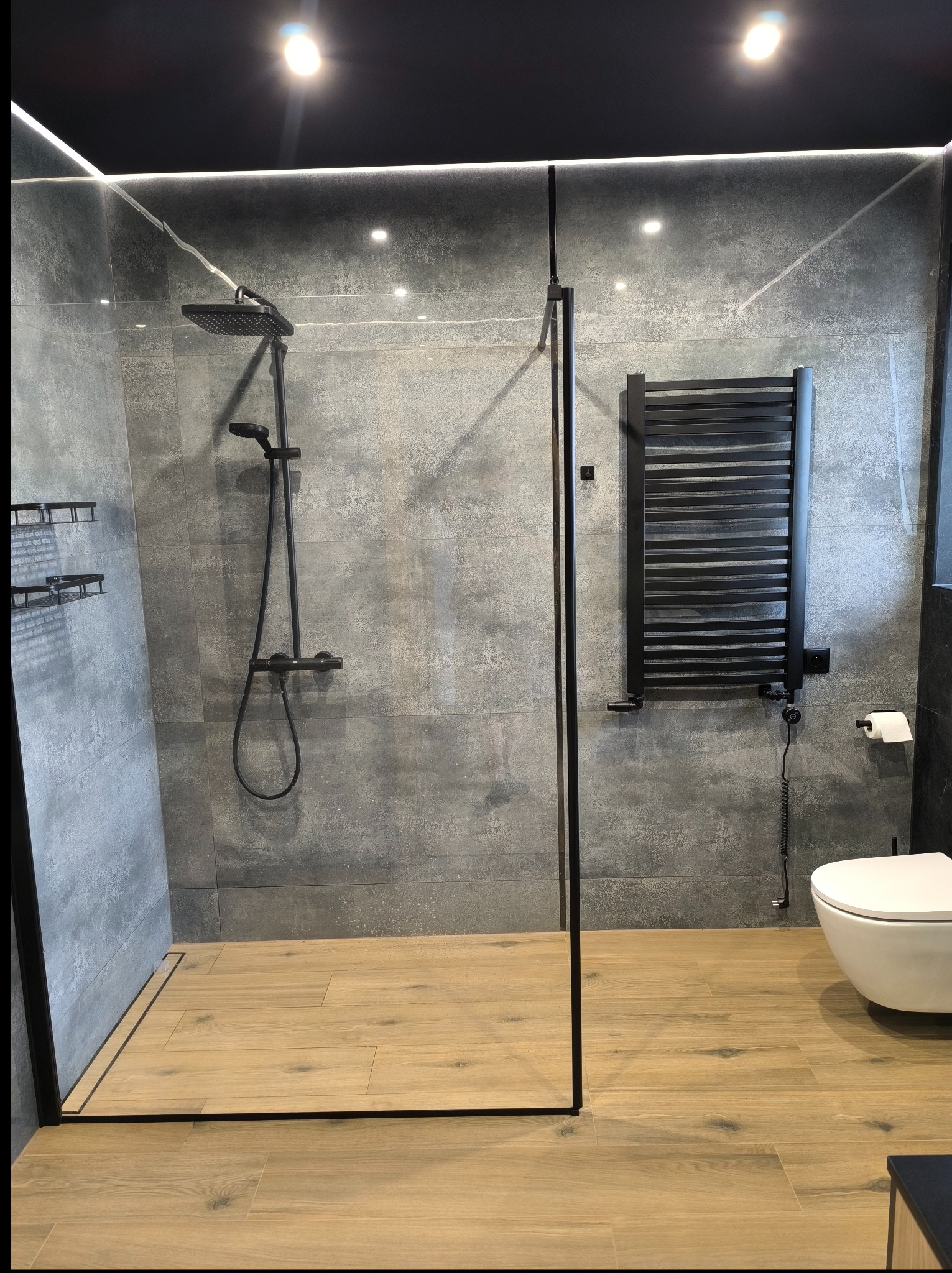 Nowoczesna łazienka z kabiną typu walk-in, szarymi płytkami imitującymi beton, drewnopodobną podłogą i czarnymi akcentami: grzejnik, armatura.