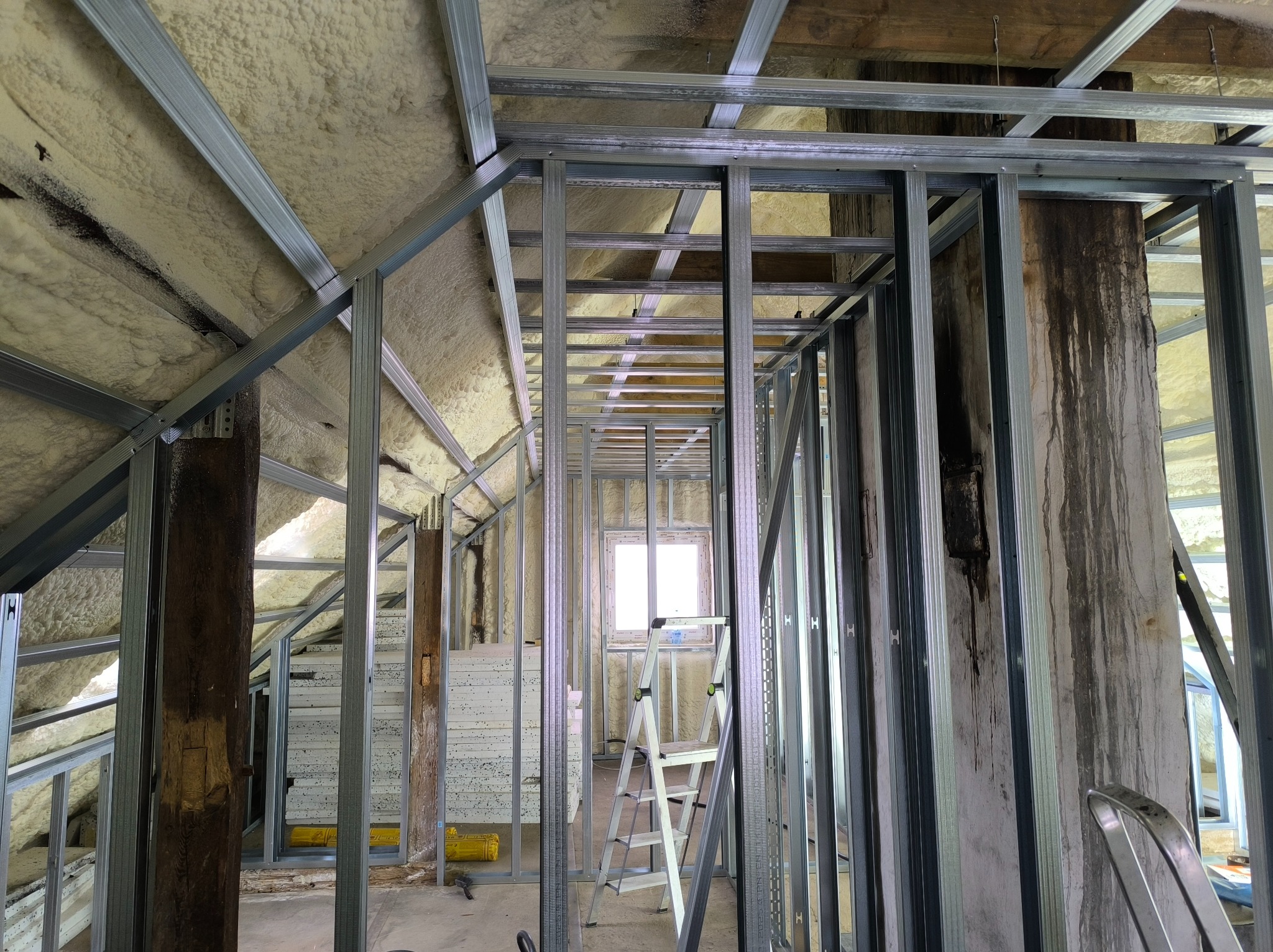 Szkielet ścianki działowej z profili stalowych w poddaszu, ocieplenie pianą PUR, drabina aluminiowa, okno w tle. Widoczne drewniane elementy konstrukcyjne.