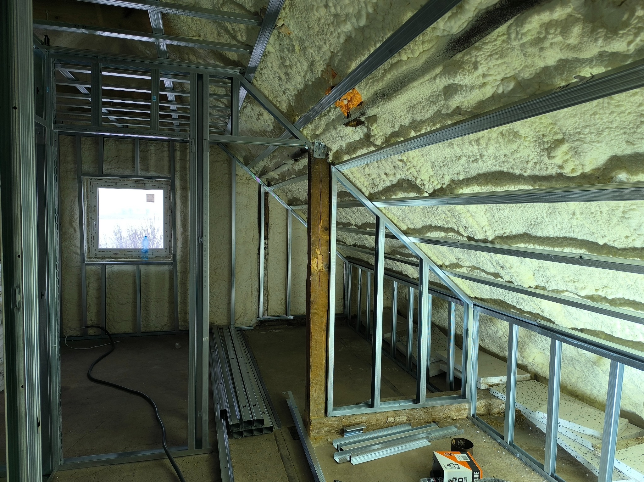 Poddasze w trakcie adaptacji: metalowa konstrukcja szkieletowa ścian działowych, izolacja pianą PUR na skosach dachu, okno. Widoczne profile i narzędzia.