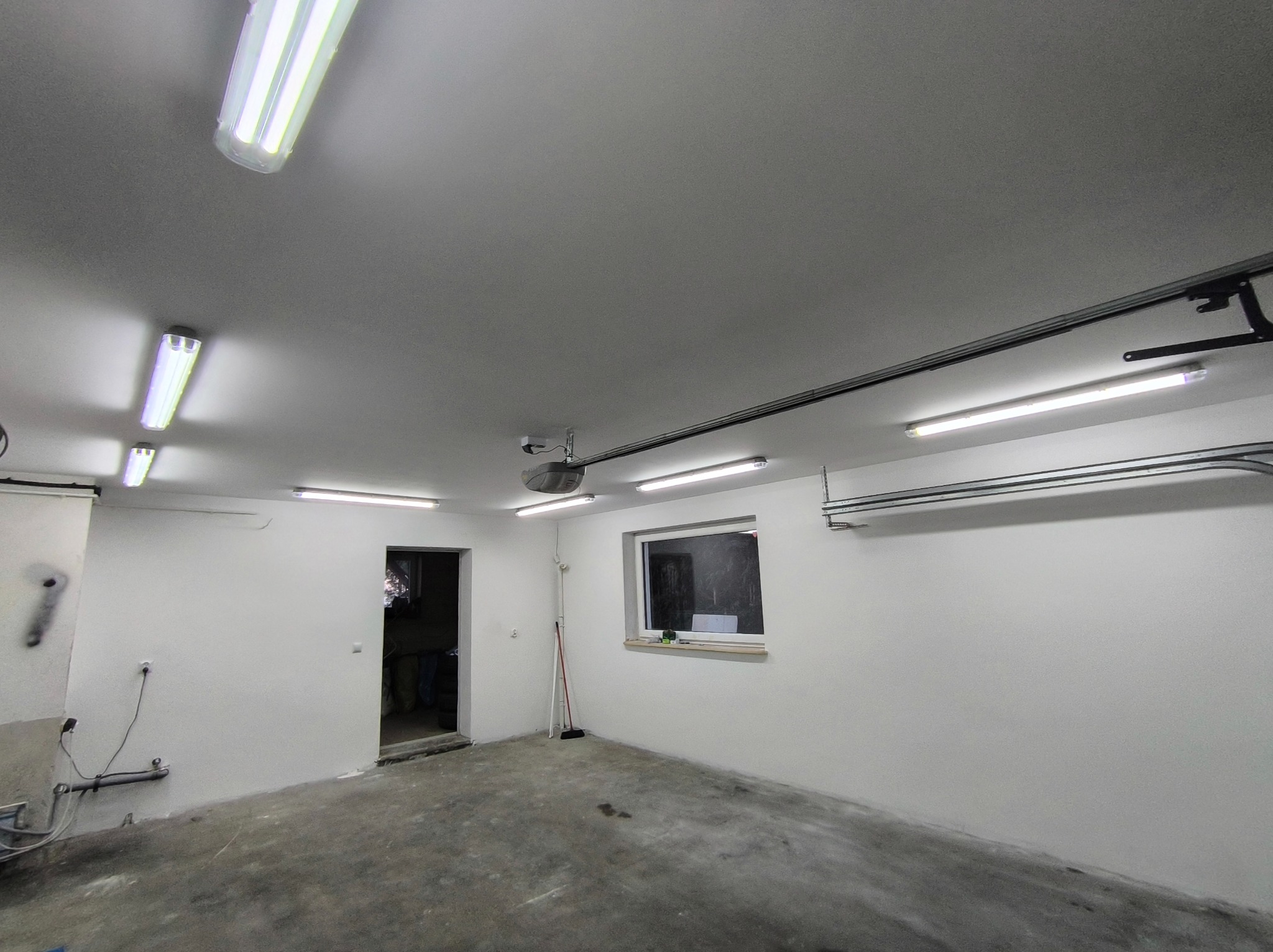 Białe, surowe wnętrze garażu z oknem i oświetleniem LED. Widoczne ślady po remoncie na podłodze. Minimalistyczny styl, surowy beton i świeżo pomalowane ściany.