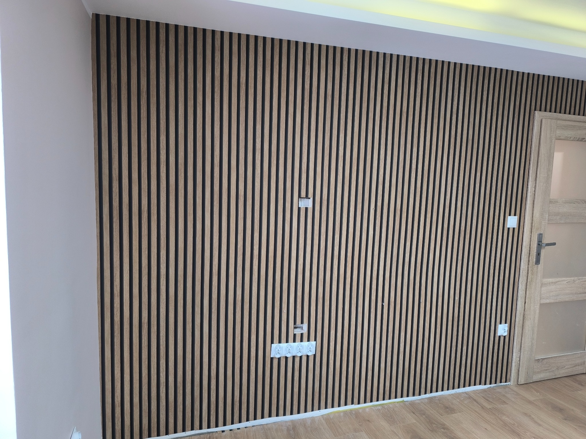 Ściana wykończona panelami lamelowymi w odcieniach drewna, z widocznymi gniazdkami elektrycznymi i fragmentem drzwi po prawej stronie. Nowoczesny design wnętrza.