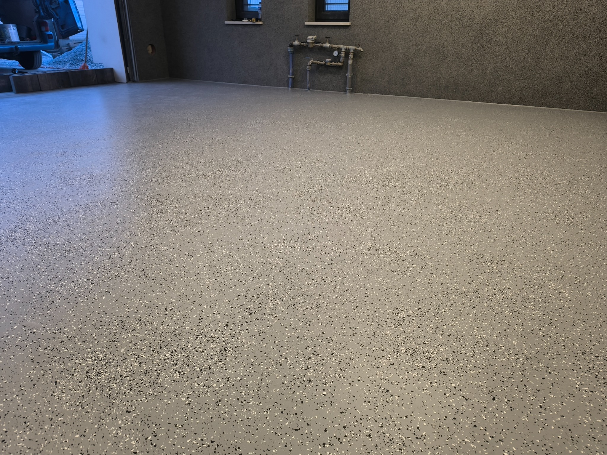 Jasnoszara posadzka żywiczna z czarnymi i białymi drobinkami w garażu. Widoczne instalacje na ścianie i częściowo otwarte drzwi garażowe z boku.